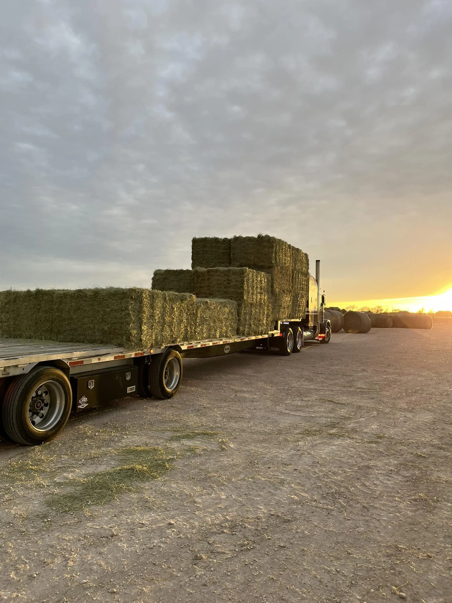 A truck carrying hay bales parked on a farm field at sunset, with more hay bales in the background and a cloudy sky overhead.