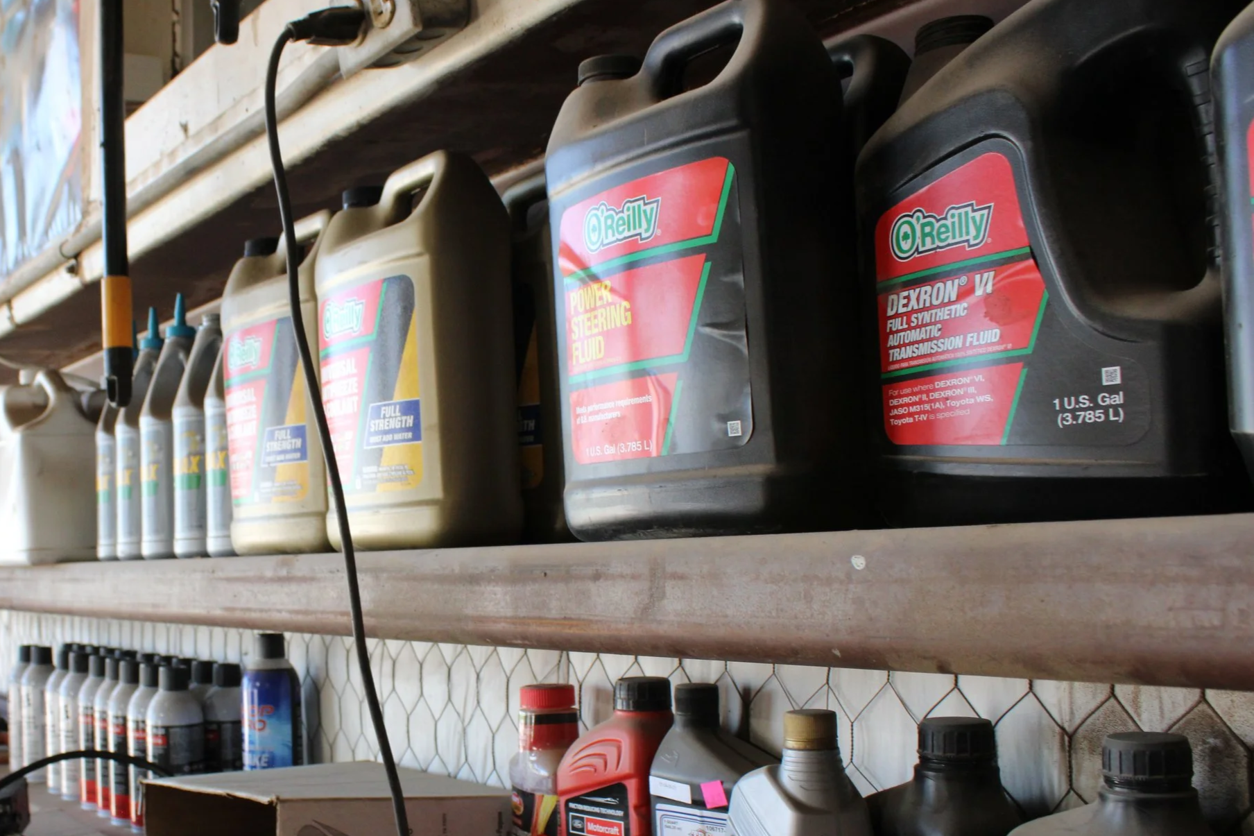 Shelves containing various containers of automotive fluids, including transmission fluid, power steering fluid, motor oil, and other products. The containers are in different sizes and colors, with some labeled with brand and product information.