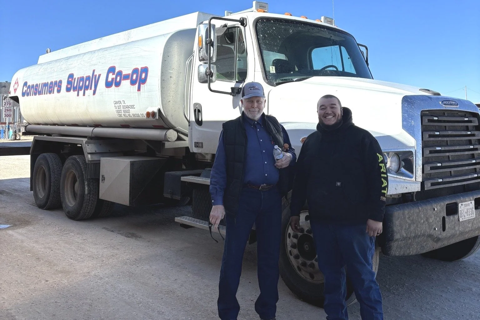 Two men standing in front of a large white fuel truck with 'Consumer Supply Co-op' written on the side, outdoors under clear blue sky.