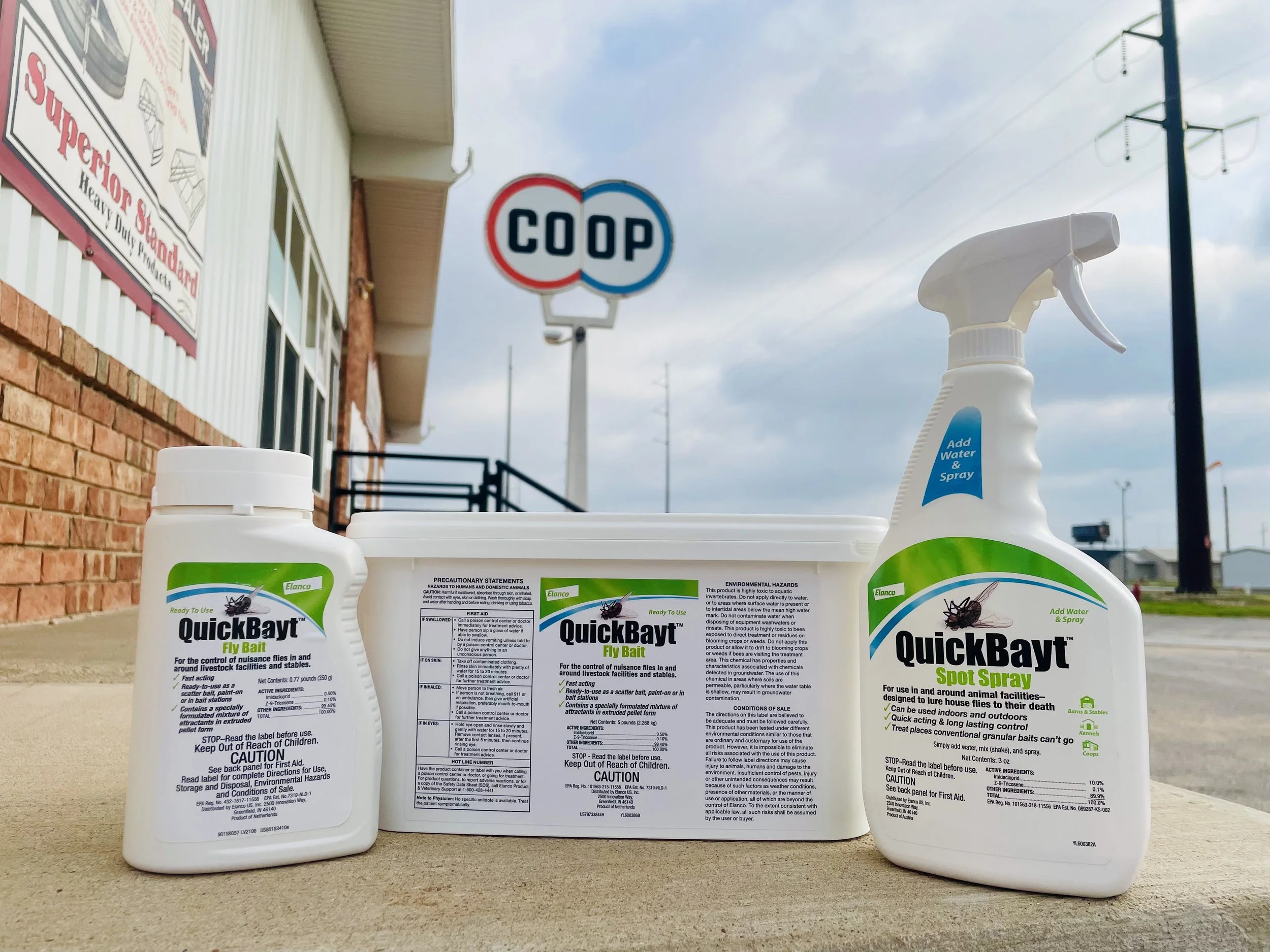 Three bottles of QuickBayt insect bait spray placed outdoors on a surface, with a grocery store in the background and a cloudy sky above.