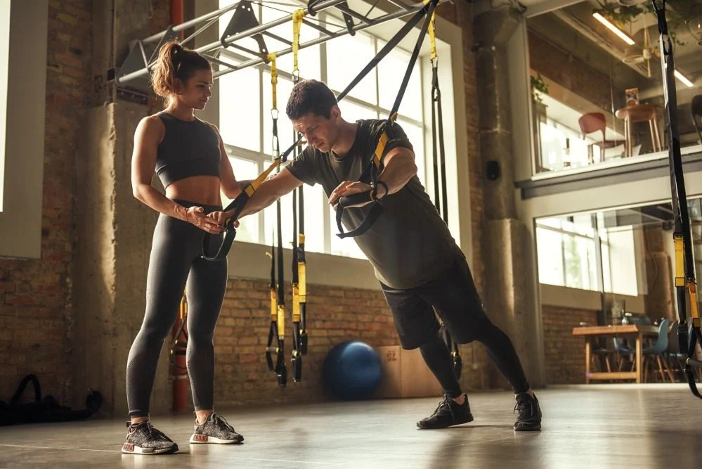 A personal trainer assists a woman with suspension training in a gym.