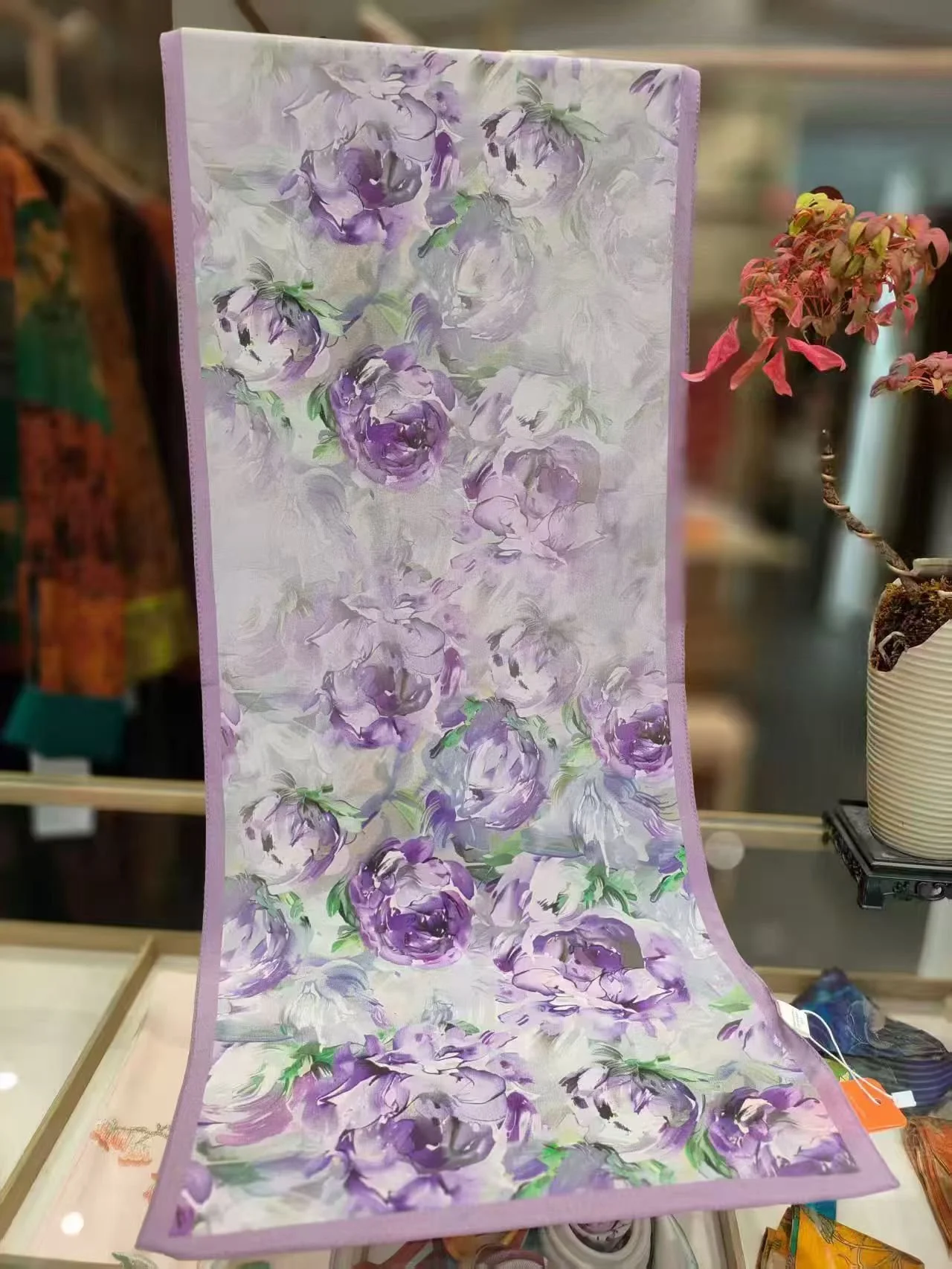 A luxurious Mulberry silk scarf featuring an elegant purple and green floral pattern, gracefully draped on a white boutique mannequin in a sunlit setting. This represents Tongbao’s product concept of bringing heritage silk into contemporary fashion.
