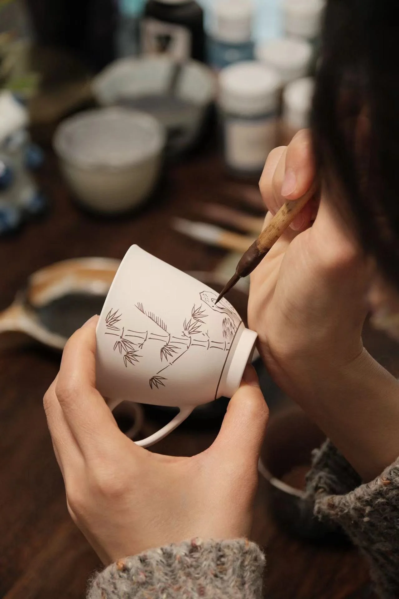 A detailed close-up of an artisan's hands meticulously hand-painting a traditional bamboo design onto a fine white porcelain cup, showcasing the centuries-old Jingdezhen blue and white porcelain craftsmanship for Tongbao.