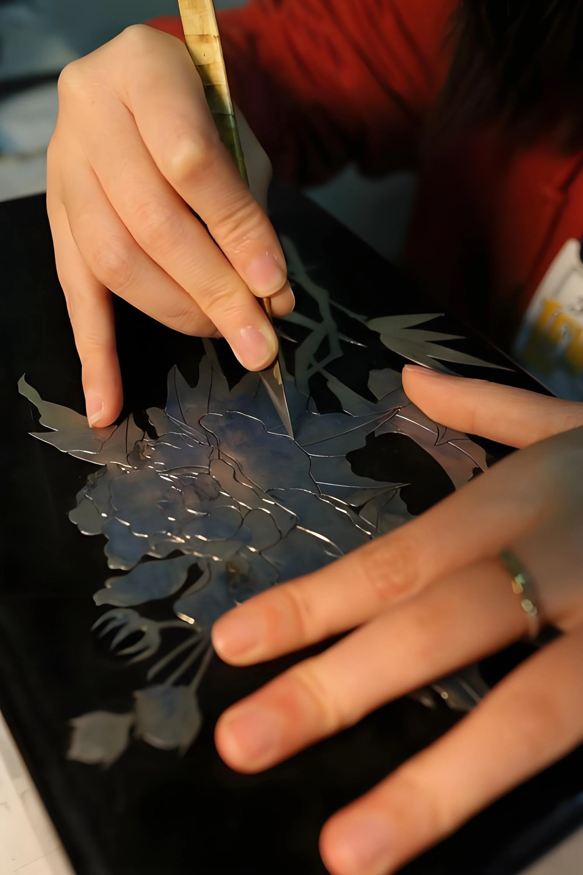 An intense editorial close-up of an artisan's focused hands using a precision tool to carve a delicate floral pattern into deep black lacquer, showcasing the patience and mastery of Tongbao’s heritage craftsmanship.