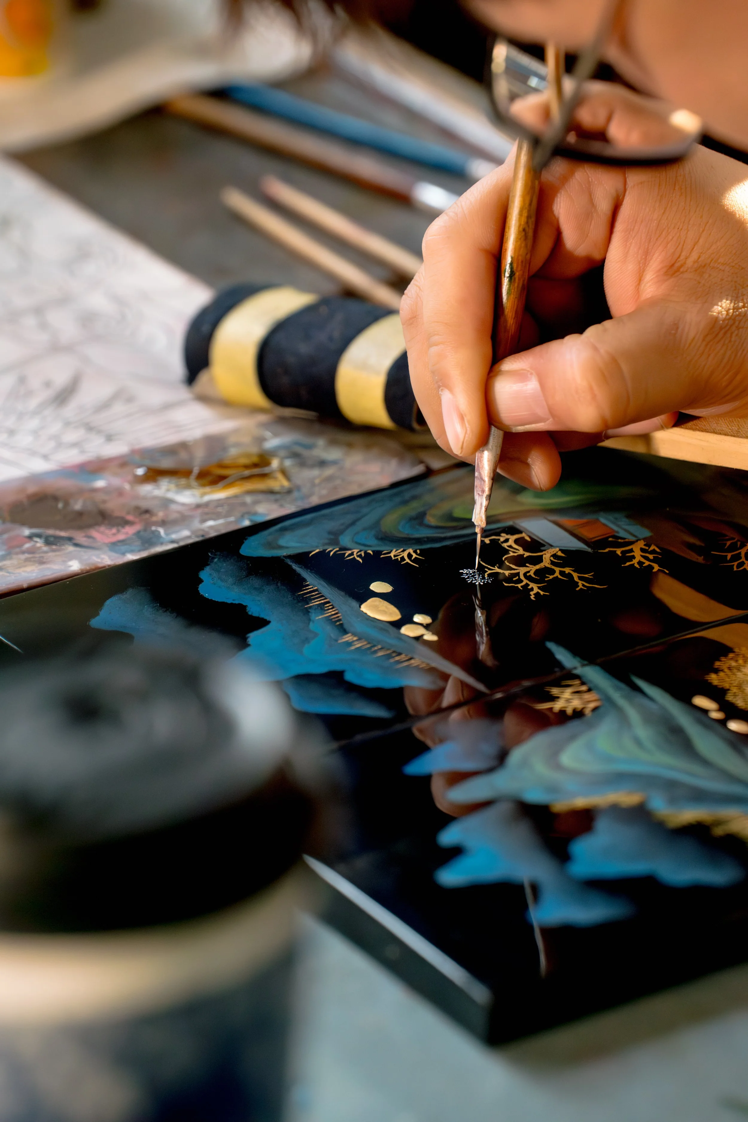 An artisan's hand uses a fine brush to delicately paint gold details onto a colourful landscape design of a handcrafted Chinese lacquerware piece, showcasing the meticulous traditional production technique for Tongbao's authentic ICH crafts.