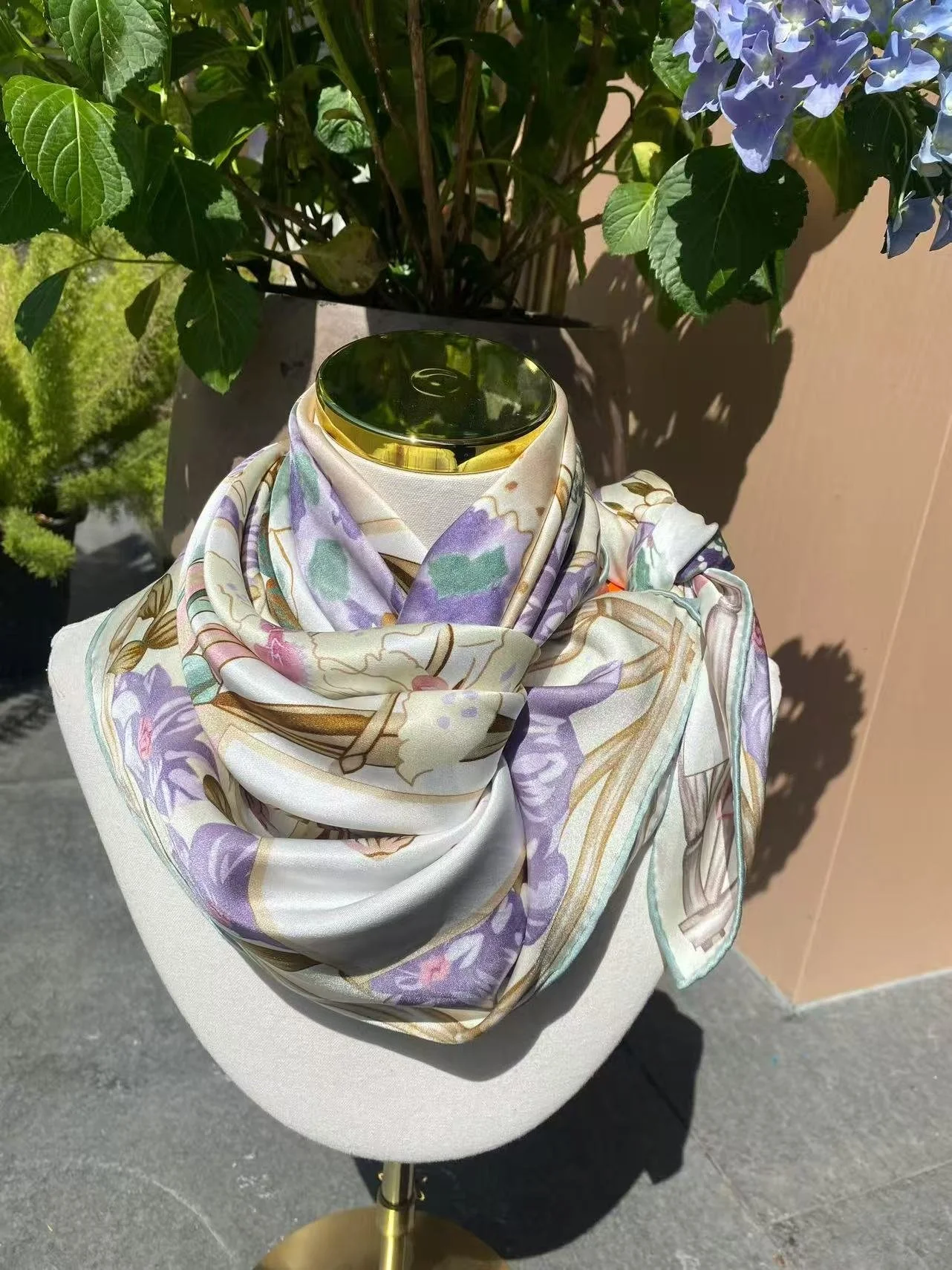 A luxurious Mulberry silk scarf featuring an elegant purple and green floral pattern, gracefully draped on a white boutique mannequin in a sunlit setting. This represents Tongbao’s product concept of bringing heritage silk into contemporary fashion.