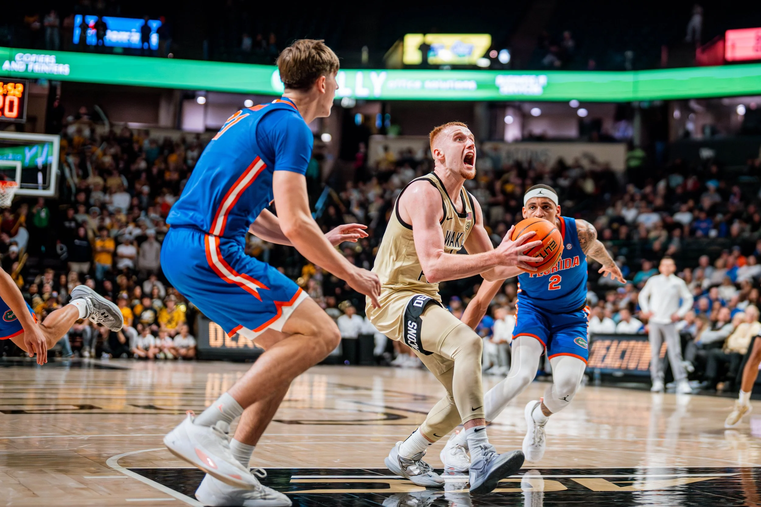 Basketball game action with three players, one in a light uniform driving toward the basket, while two players in blue uniforms defend on a court with spectators in the background.