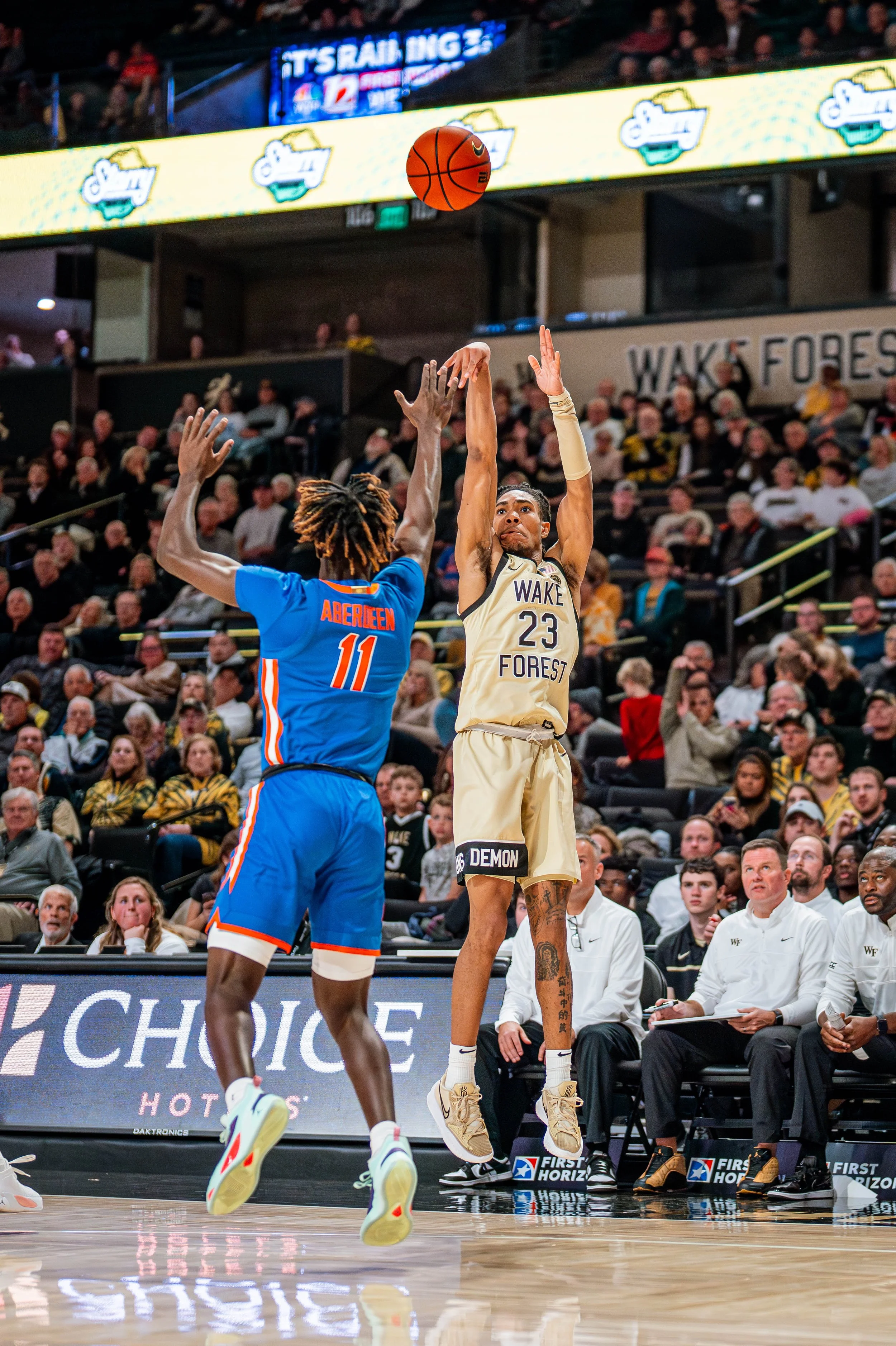 College basketball game with player from Wake Forest shooting a jump shot while being defended by a player from an opposing team in blue jersey. Crowd in the background.