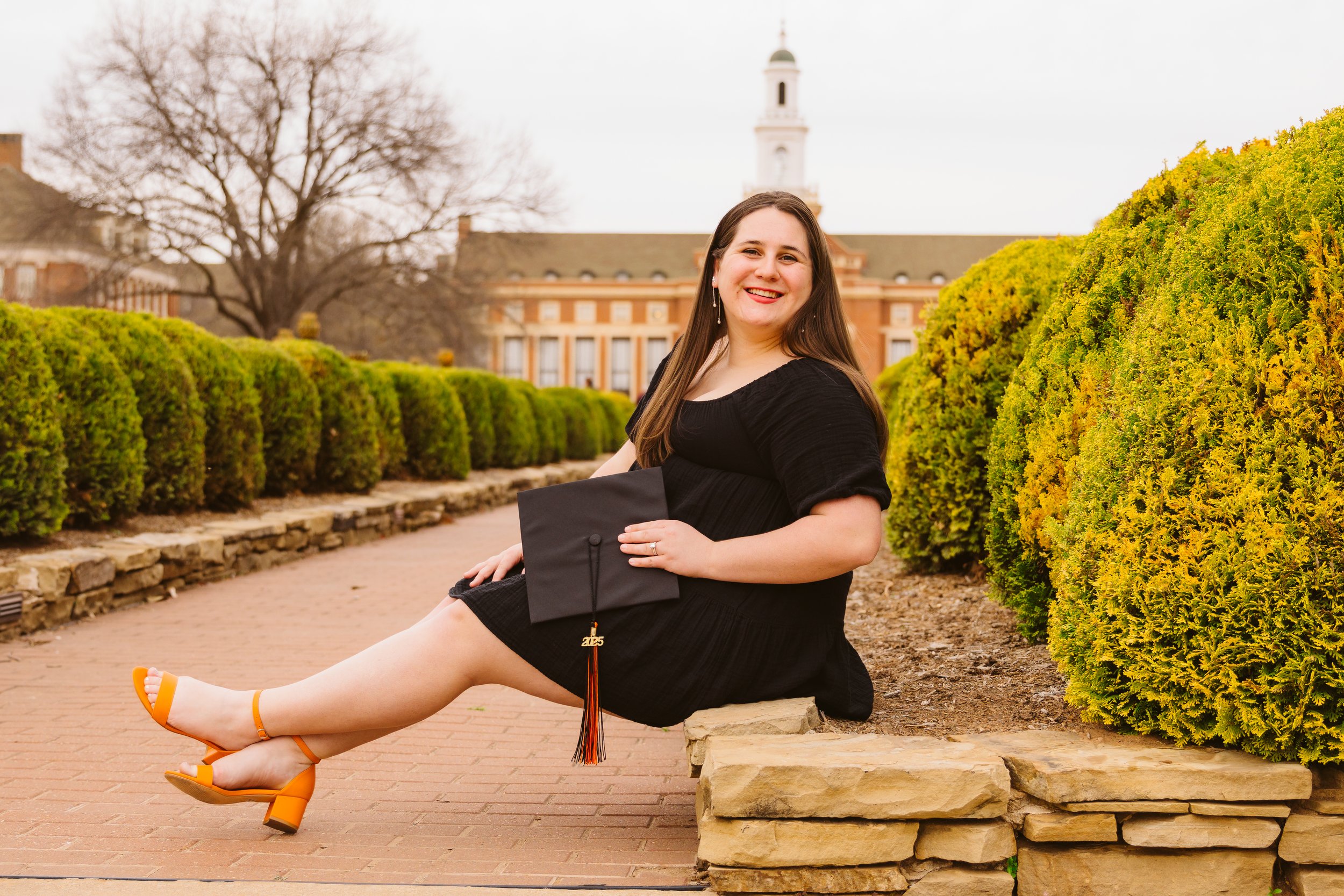 Woman in black dress sitting on stone bench holding graduation cap with '2023' tassel, surrounded by green shrubs and historic building in background.
