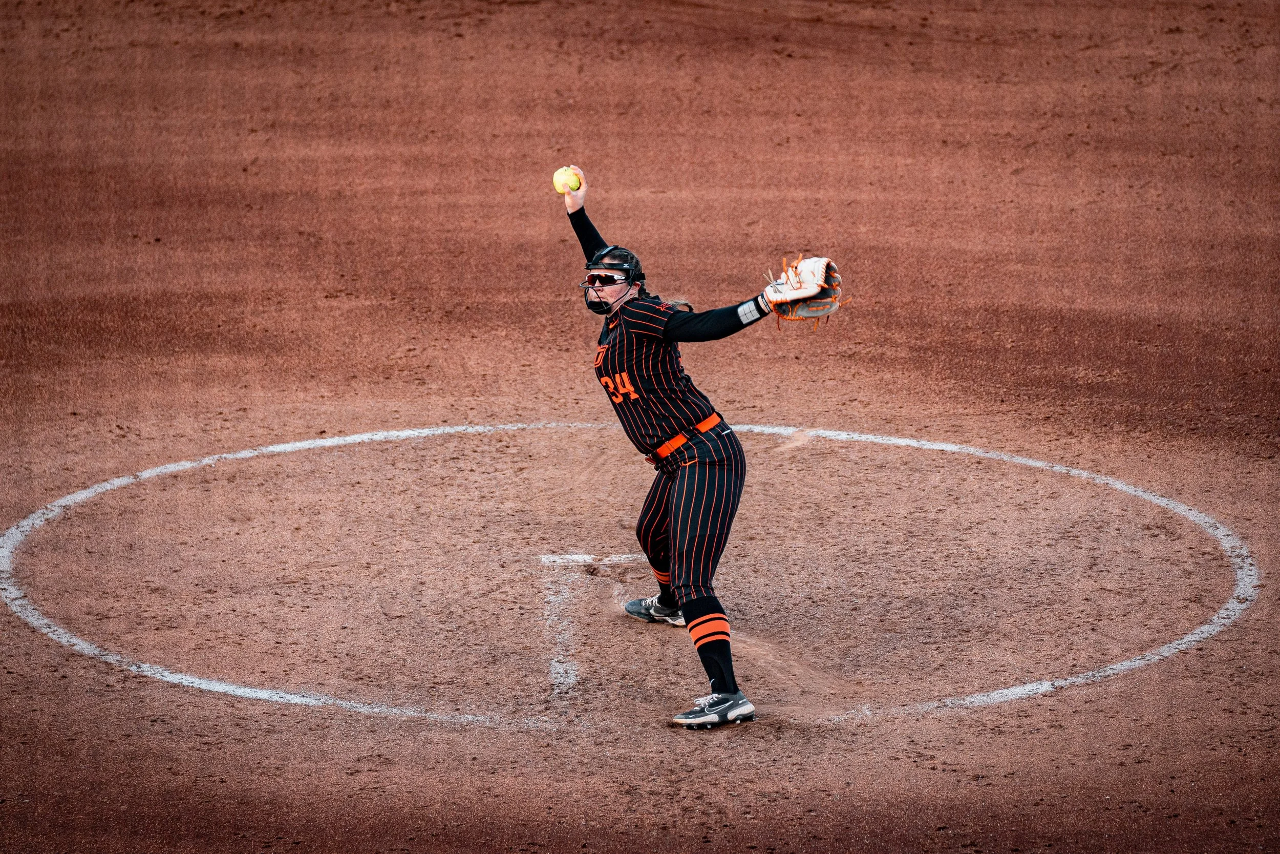 Softball player pitching on a dirt field wearing black-striped uniform.