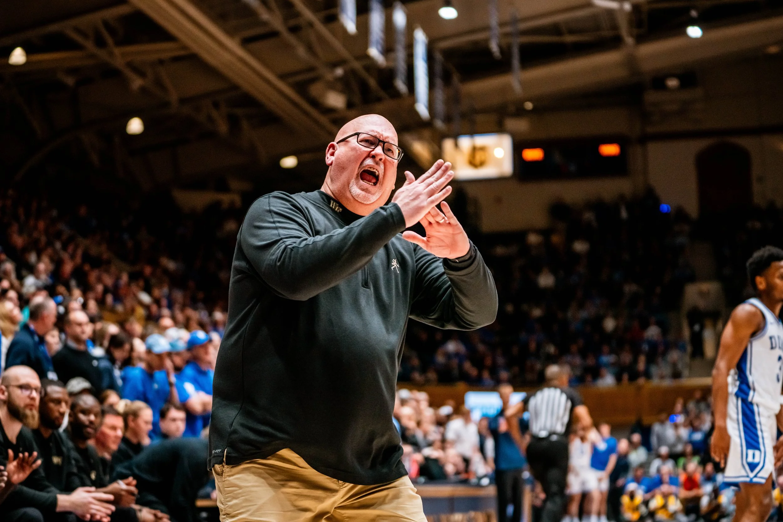 Basketball game with a coach passionately gesturing on the sideline, surrounded by fans in a crowded arena.