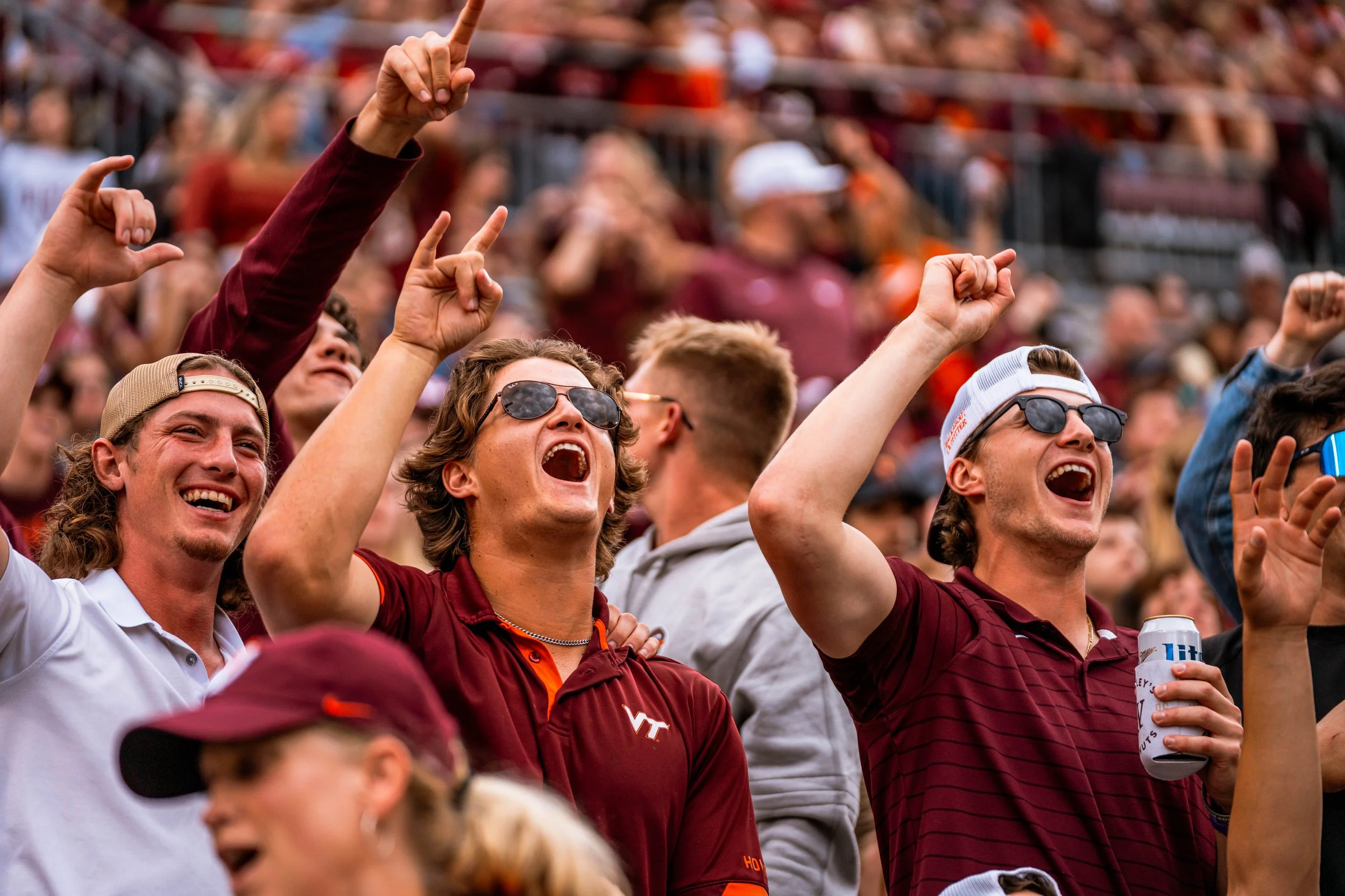 Excited crowd of sports fans cheering, wearing sunglasses and casual clothing, at an outdoor event or game.