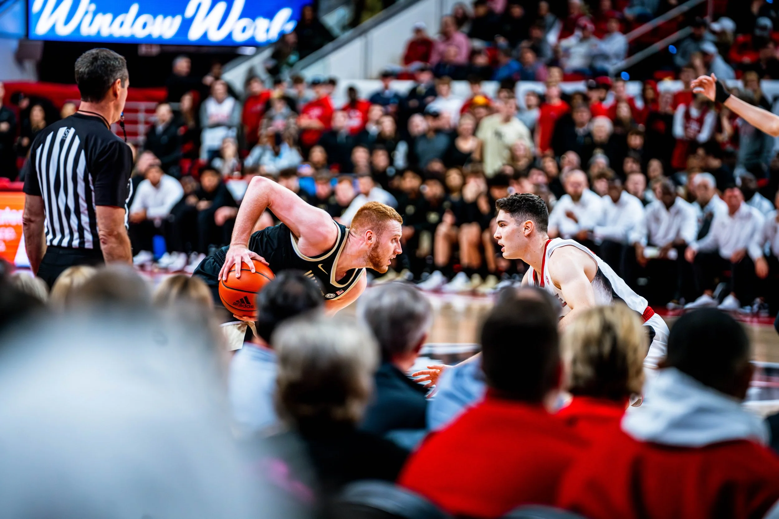Basketball player in black jersey dribbling, facing a defender in white jersey, during a game with spectators and referee in the background.
