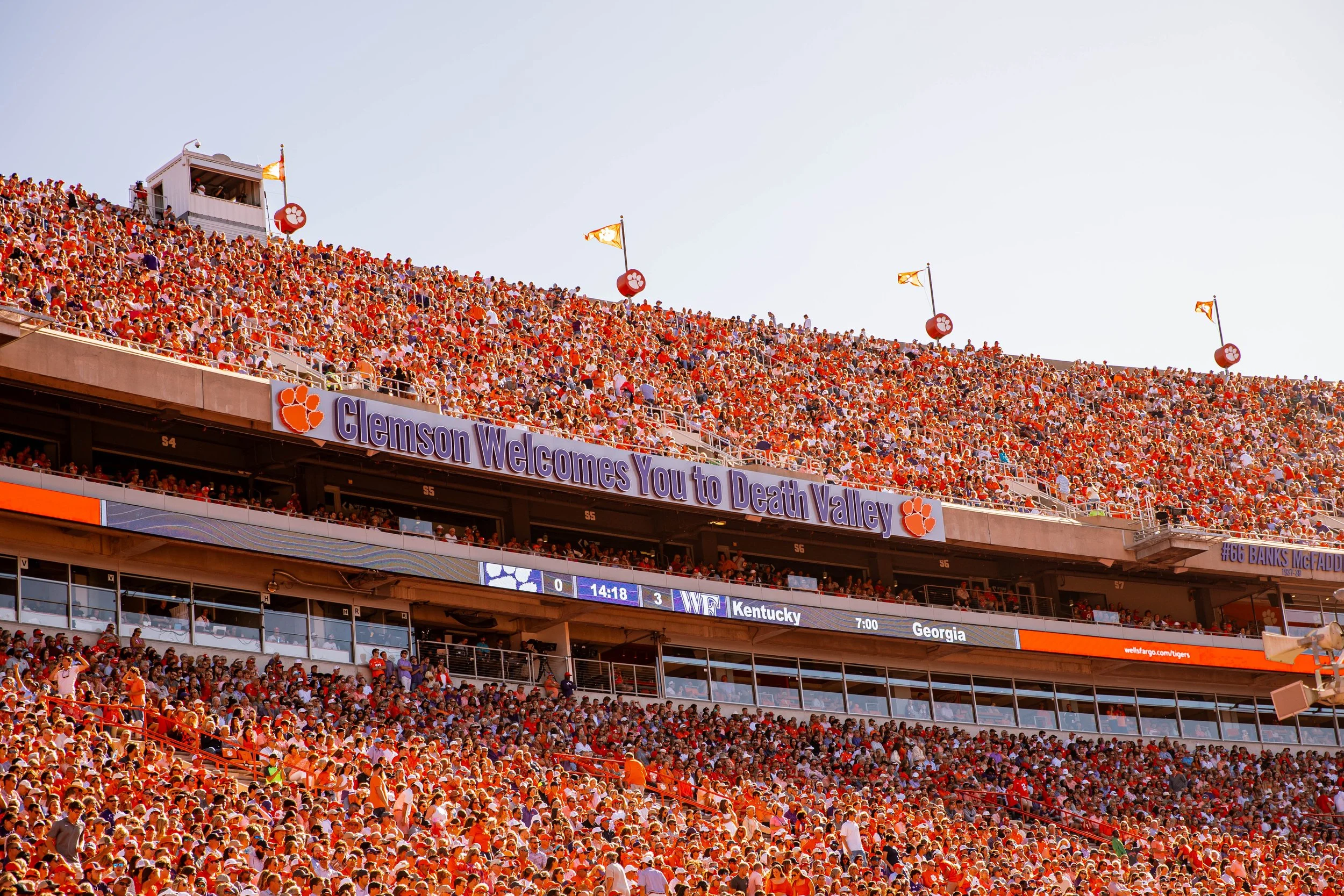 Crowded Clemson University football stadium with "Clemson Welcomes You to Death Valley" banner, scoreboard showing "Kentucky," and fans in orange.