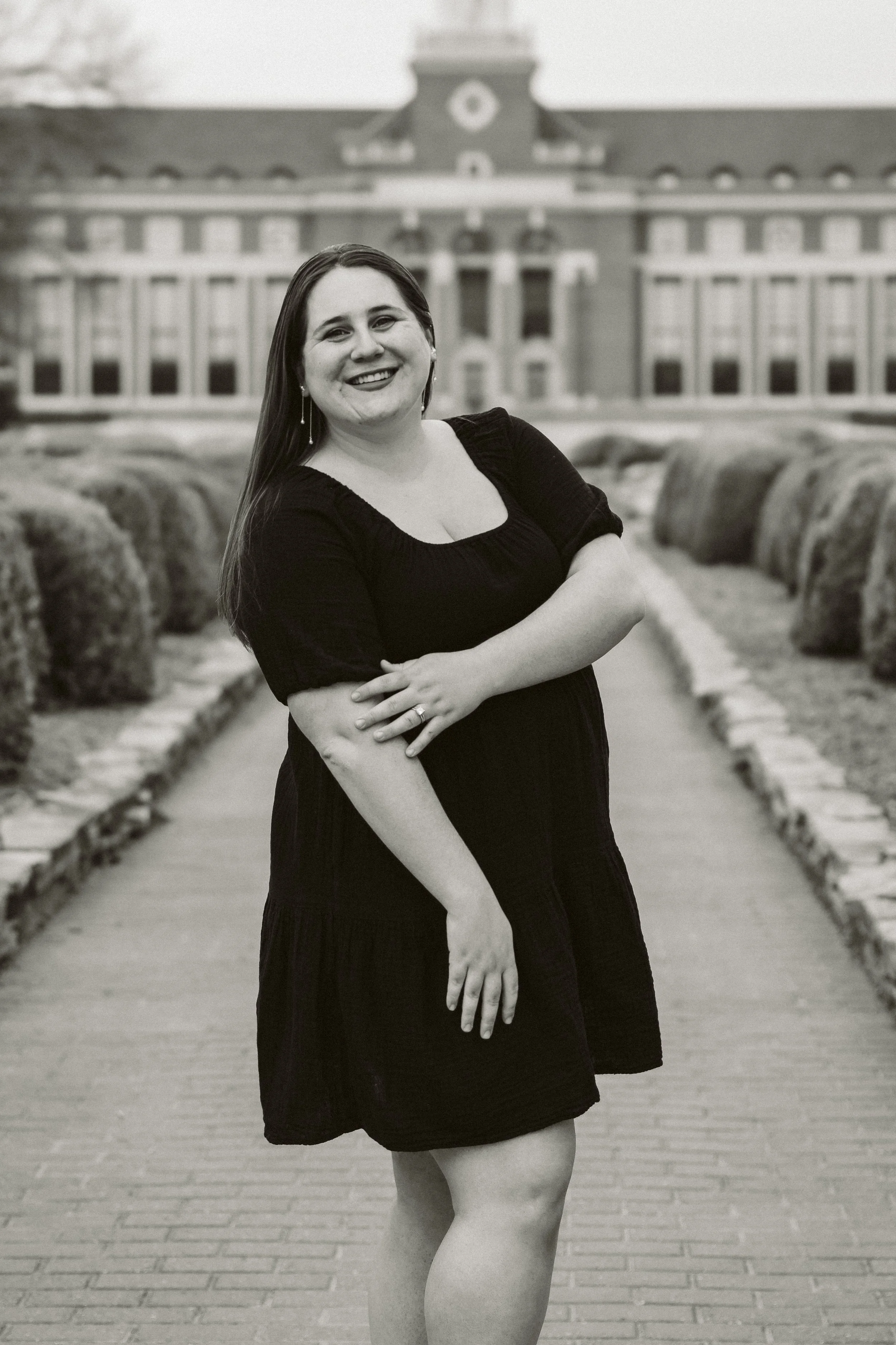 Woman in dark dress smiling in front of a large building with columns, standing on a brick path surrounded by shrubbery. Black and white photo.