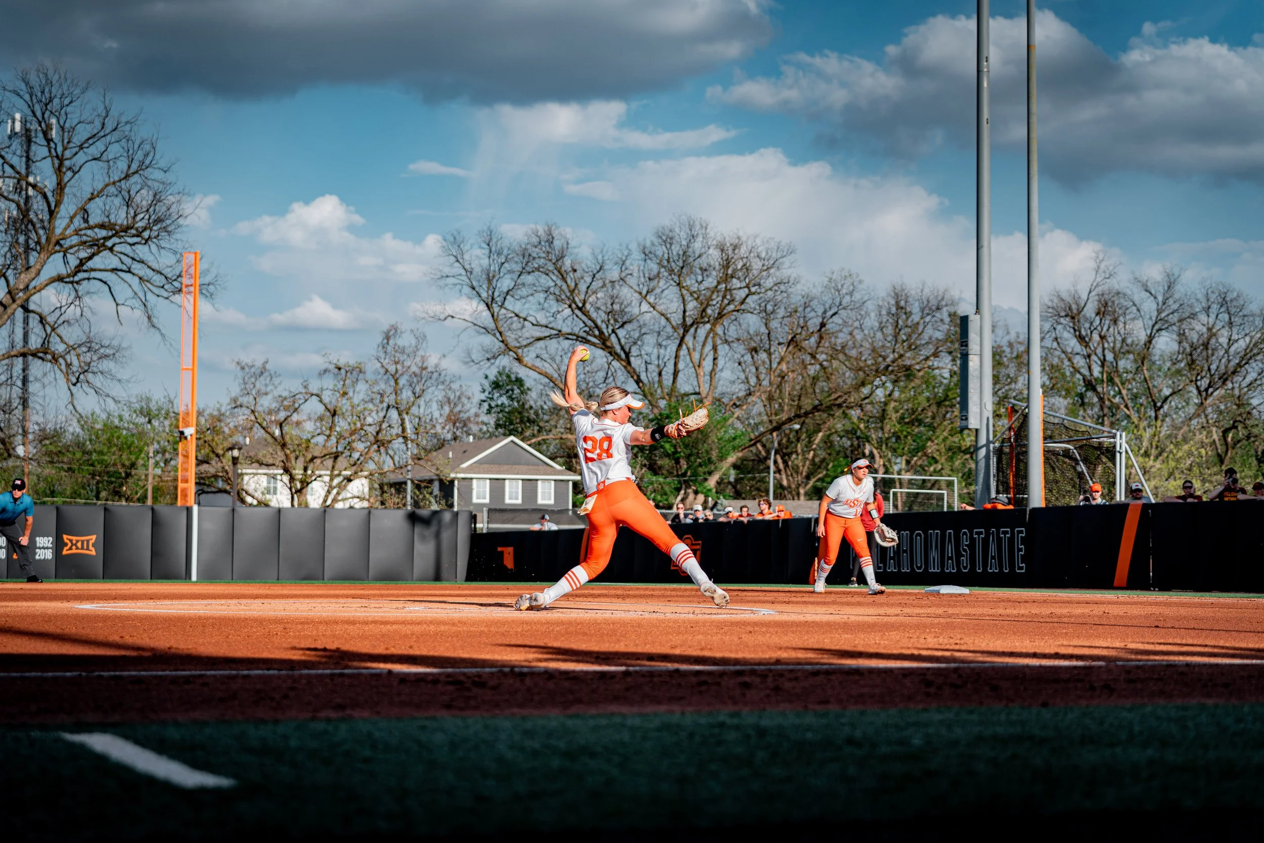 Softball pitcher in action on a field under blue skies, wearing orange and white uniform, with teammates and spectators in the background.