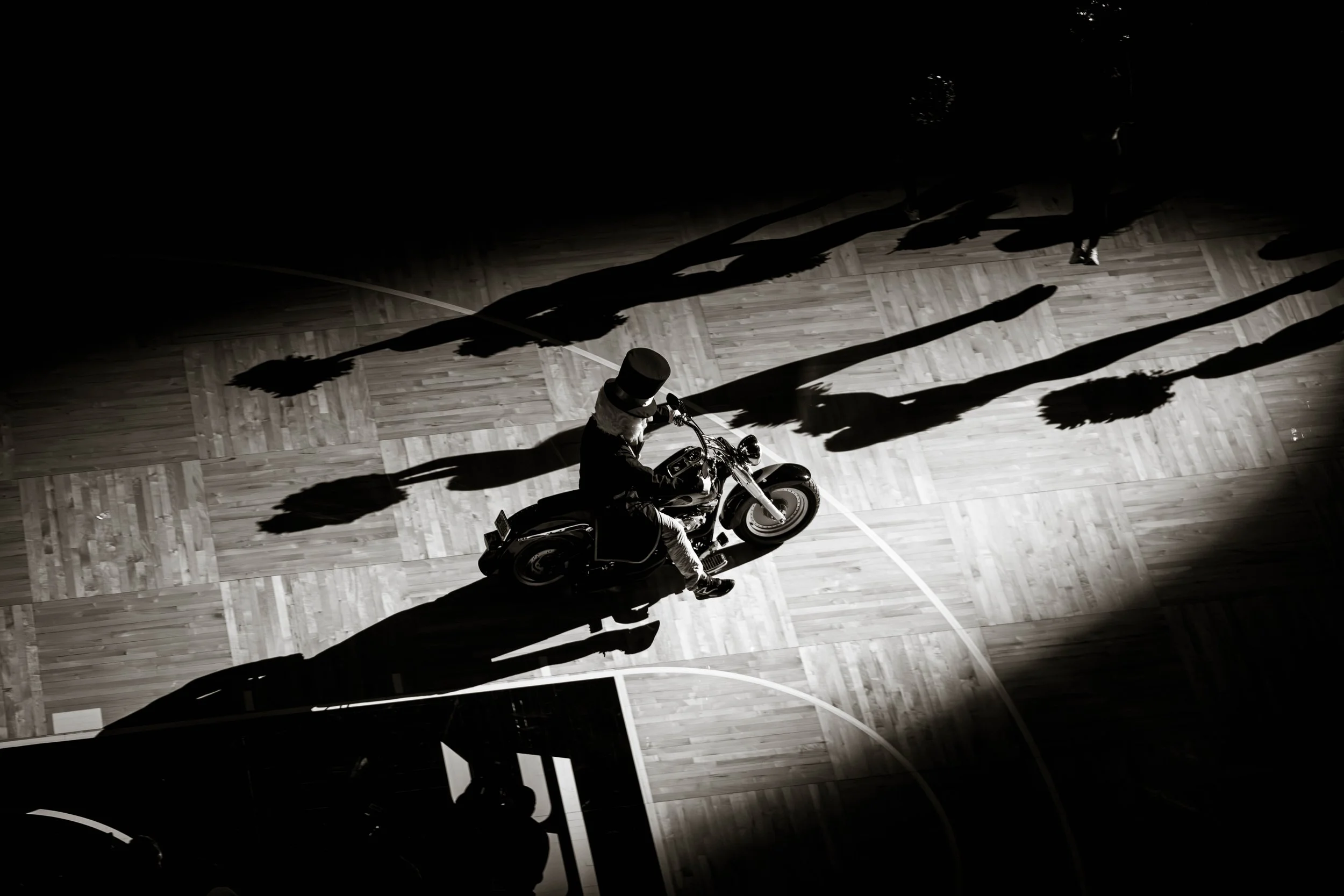 Man wearing a top hat riding a motorcycle indoors on a wooden floor with shadows of people holding pom-poms around.