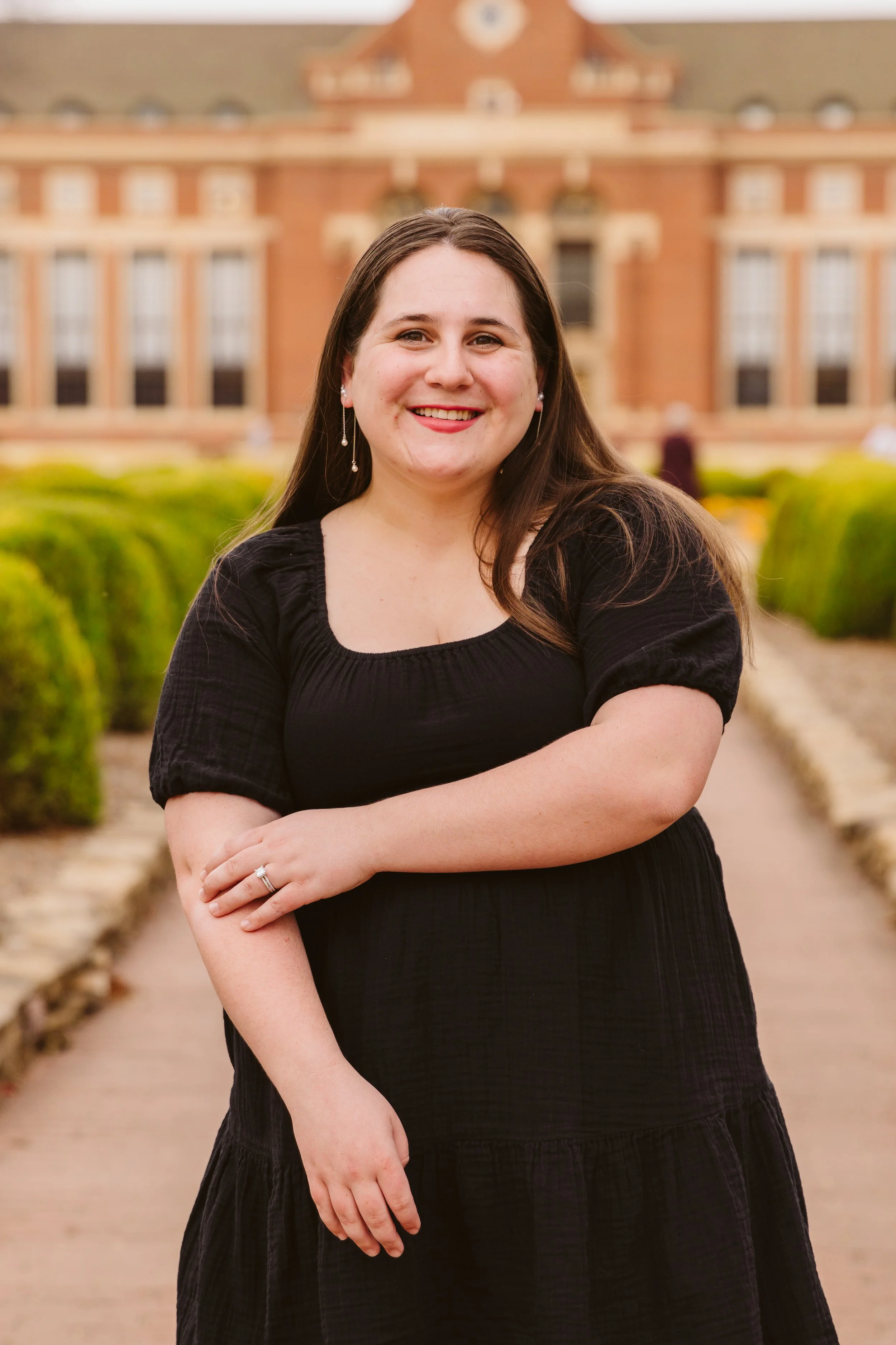 Woman in a black dress smiling outdoors with a brick building in the background.