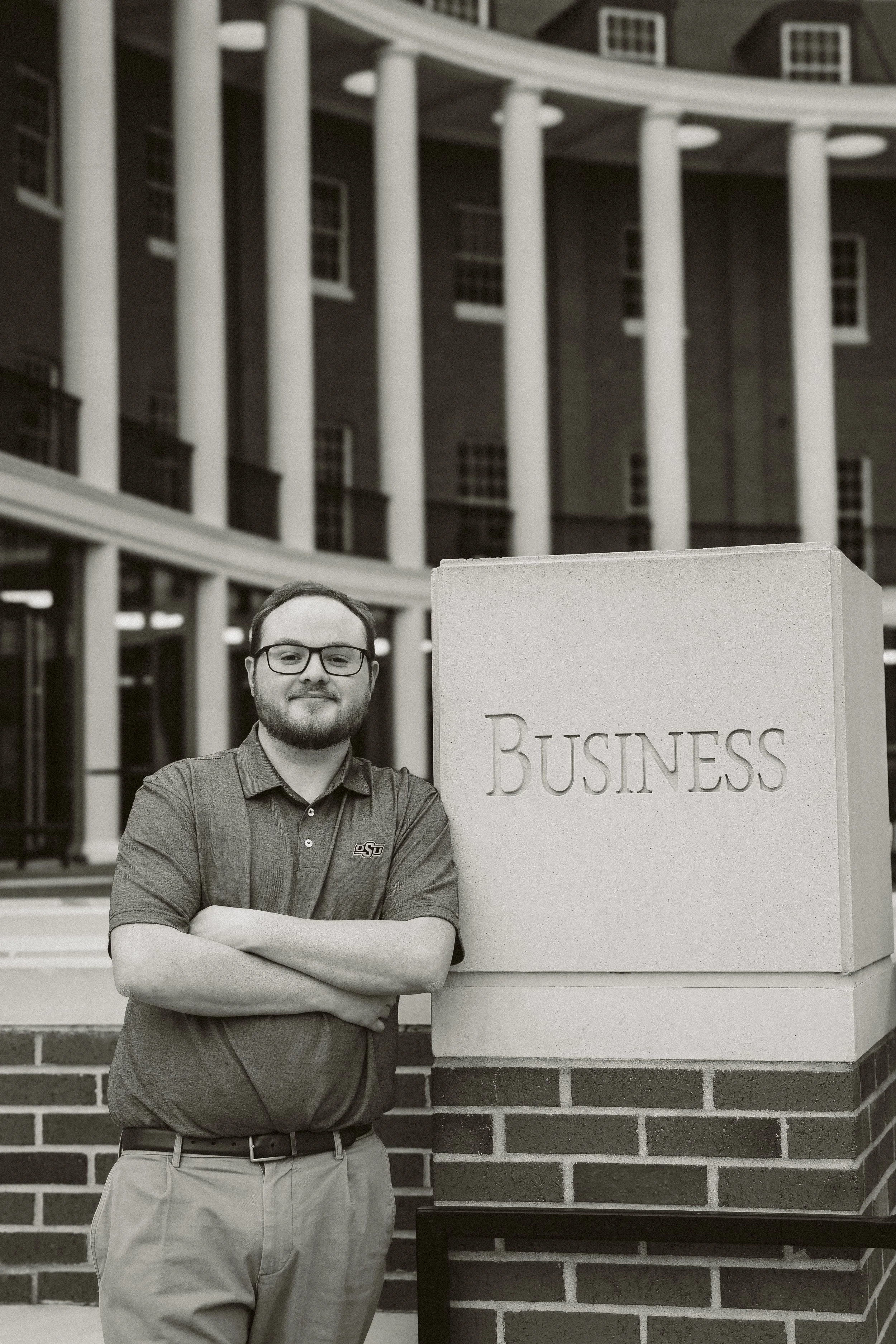 Person with glasses and a beard standing next to a sign with the word 'Business' outside a building with columns.