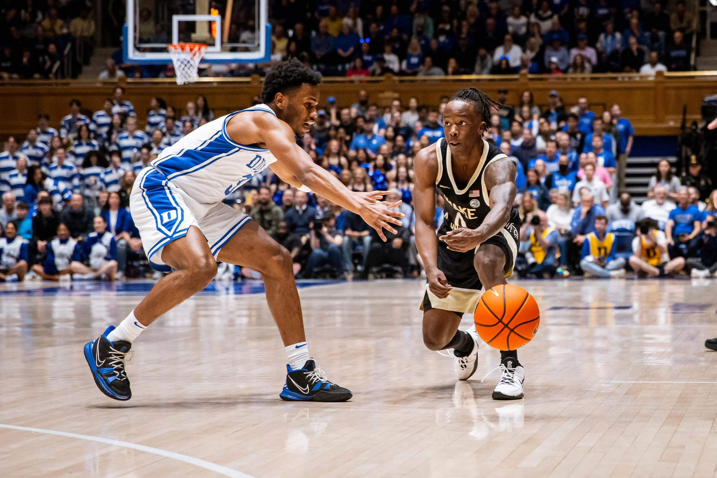 Two college basketball players from opposing teams vying for control of the basketball on a court during a game, with spectators visible in the background.