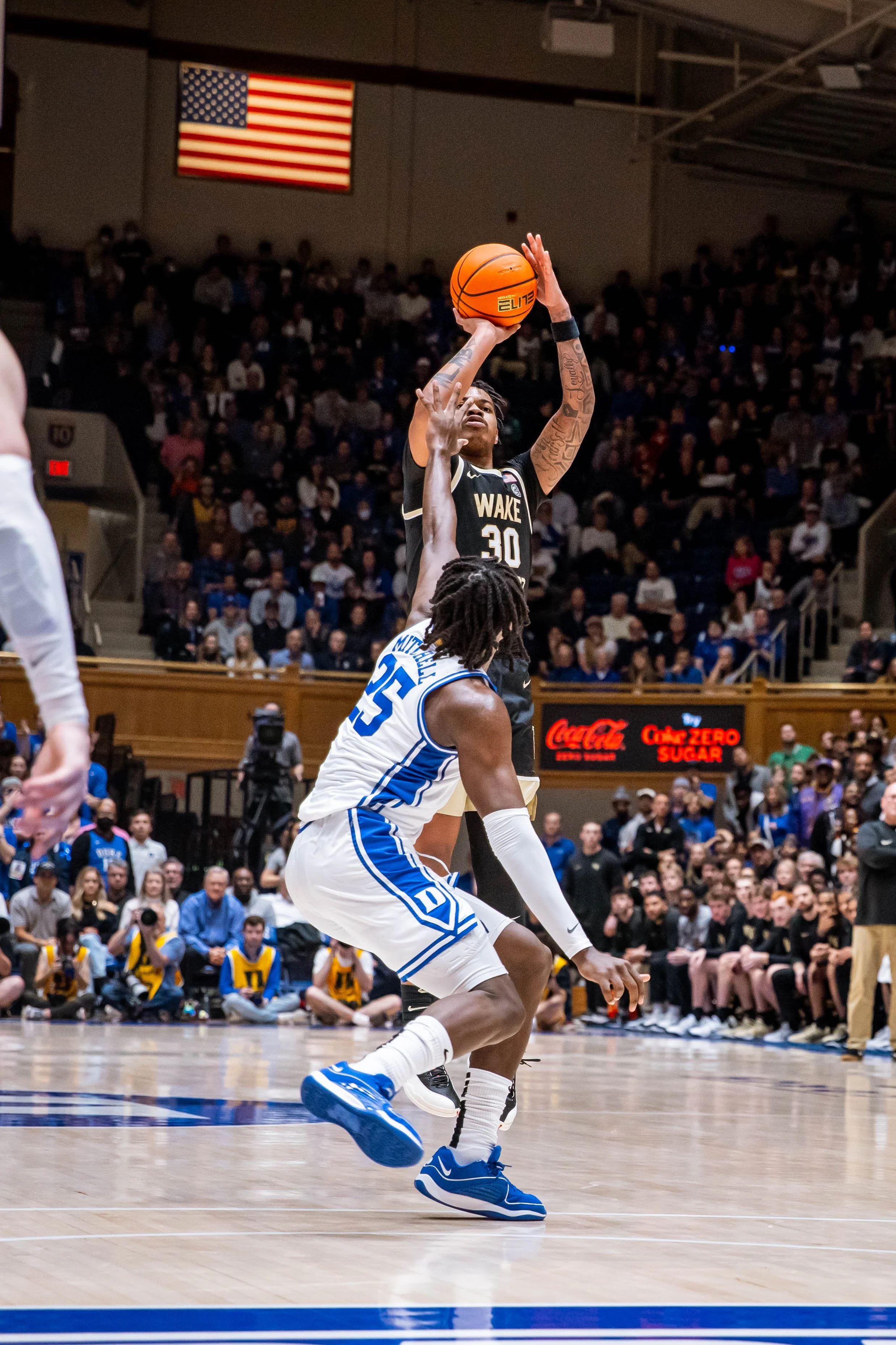 Basketball player from Wake Forest shooting over Duke defender in a game, with an American flag in the background.