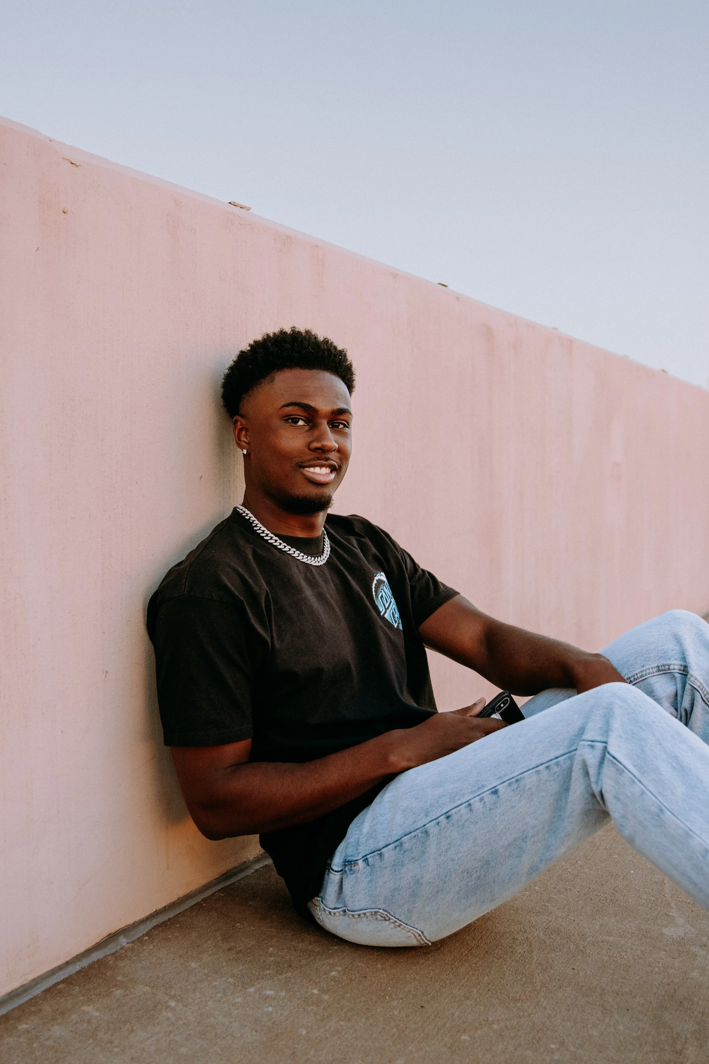 Man sitting against a pink wall wearing a black T-shirt and light blue jeans.