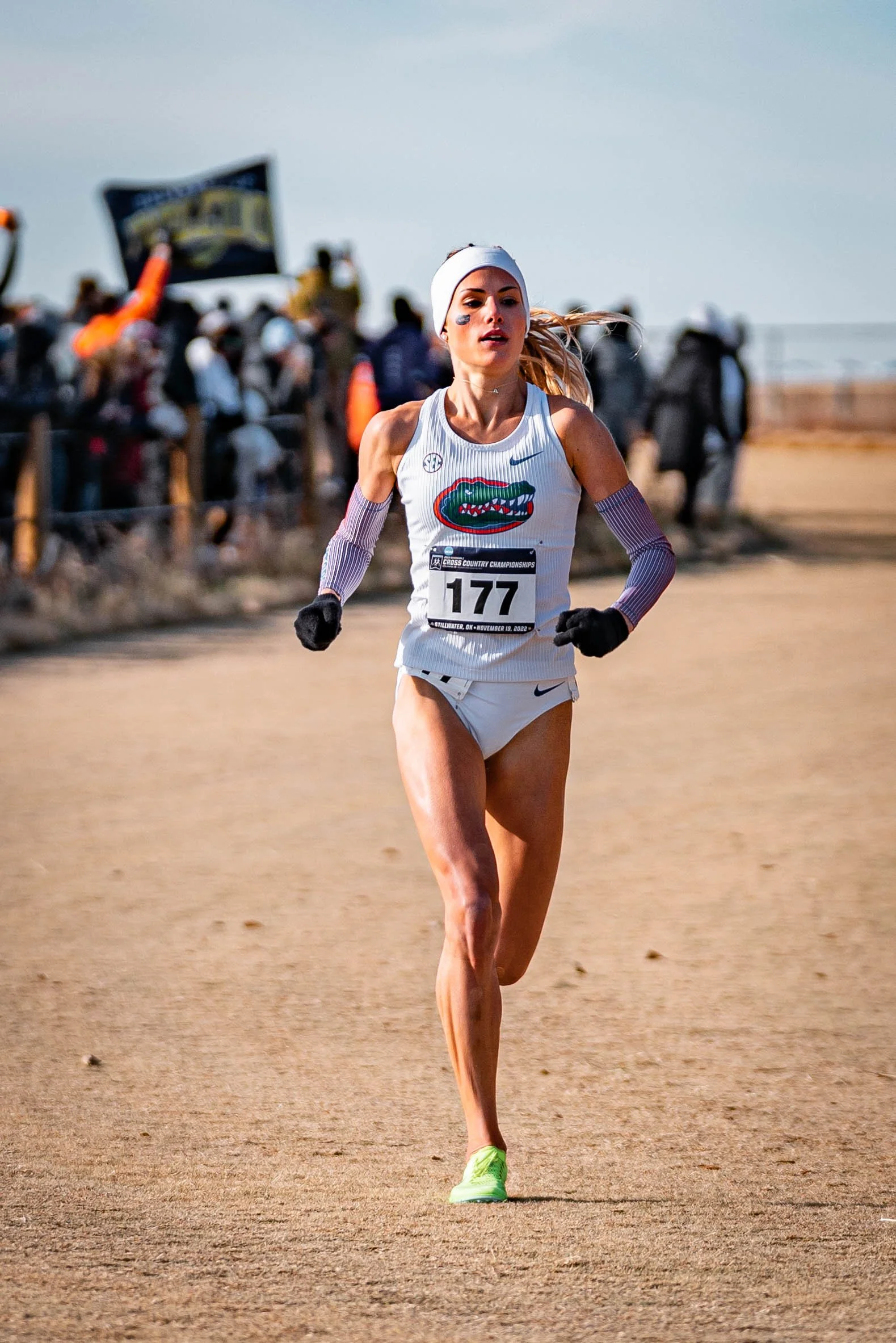 Female athlete running in a cross-country race, wearing a Florida Gators uniform with number 177, spectators and banners in the background.