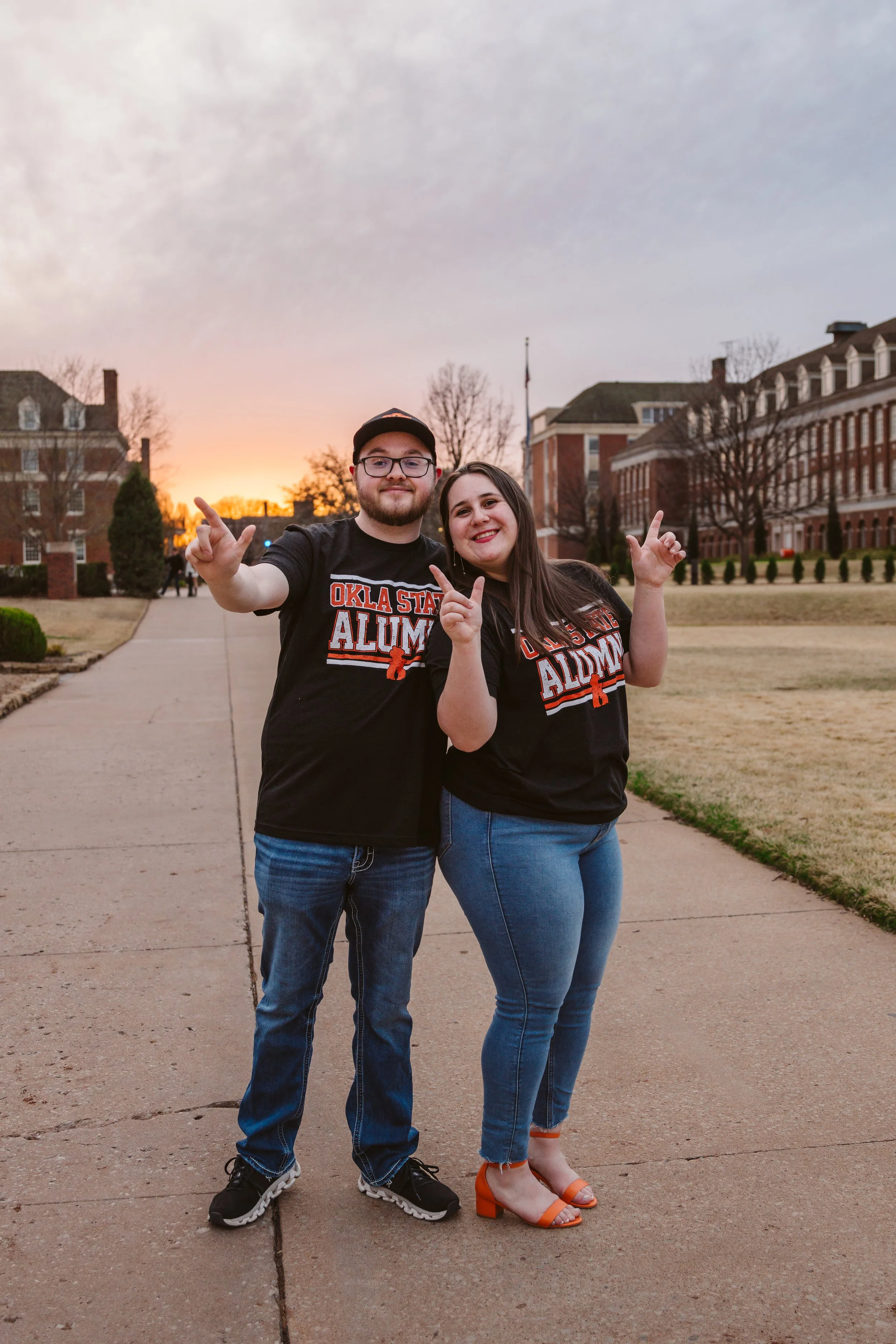 Two people wearing Oklahoma State Alumni shirts, standing on a campus sidewalk at sunset, posing with hands raised.