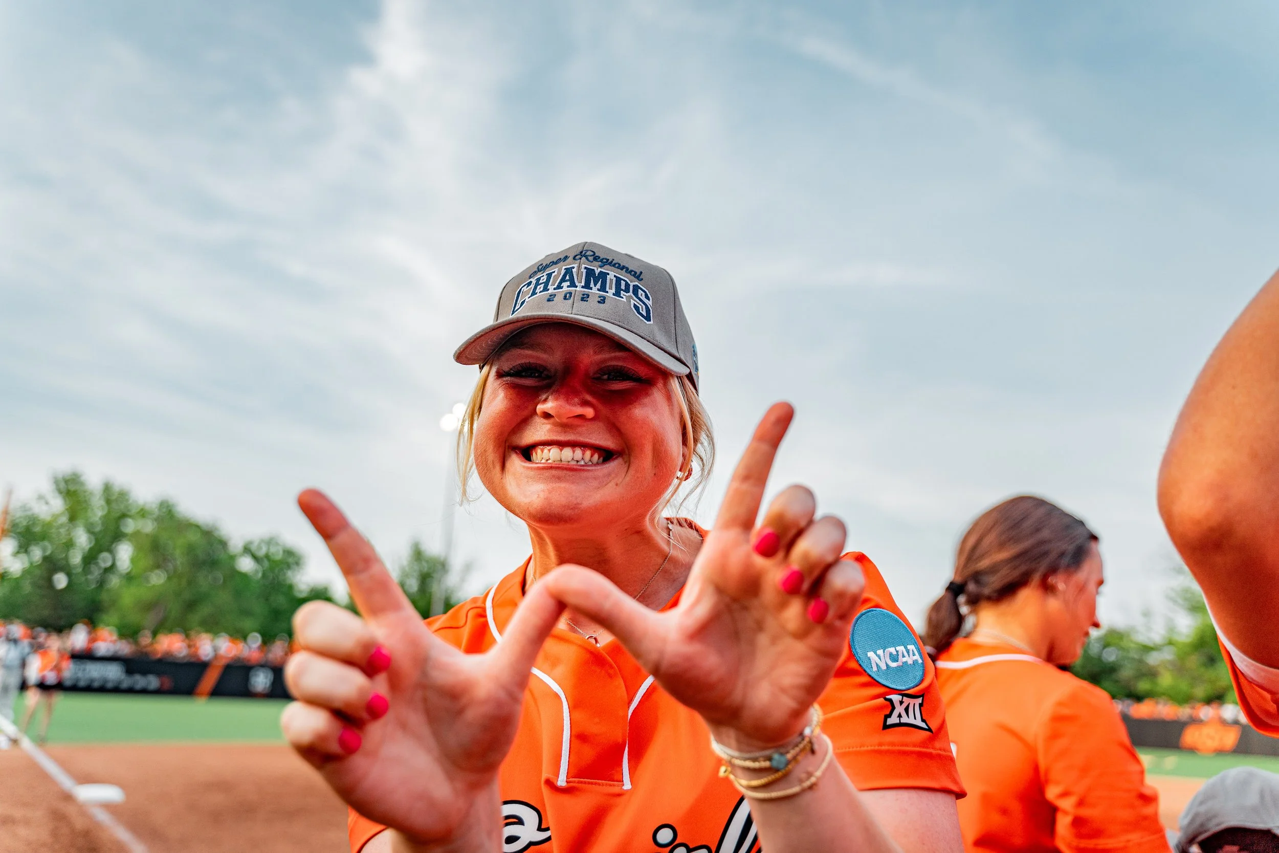 A smiling woman in an orange sports jersey and gray cap on a baseball or softball field. She is making a "W" gesture with her hands and wearing a cap that says "Champs 2023," with "NCAA" and conference patches on her sleeve. Another person in similar