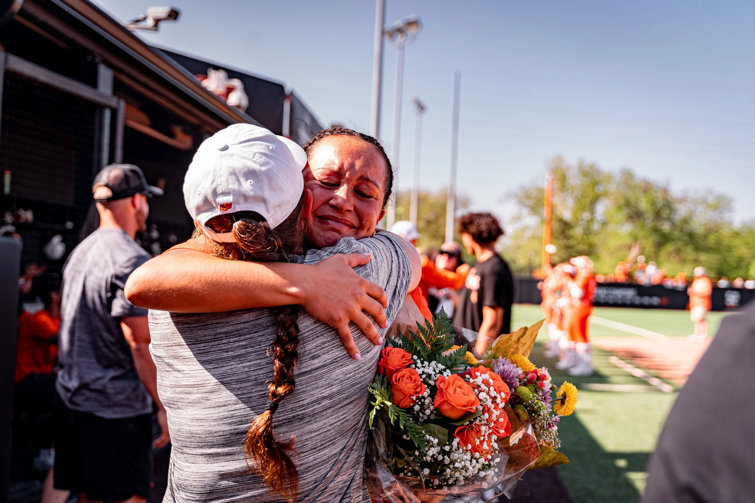 Two people embracing, one holding a bouquet of orange and white flowers, on a sports field with others in the background.