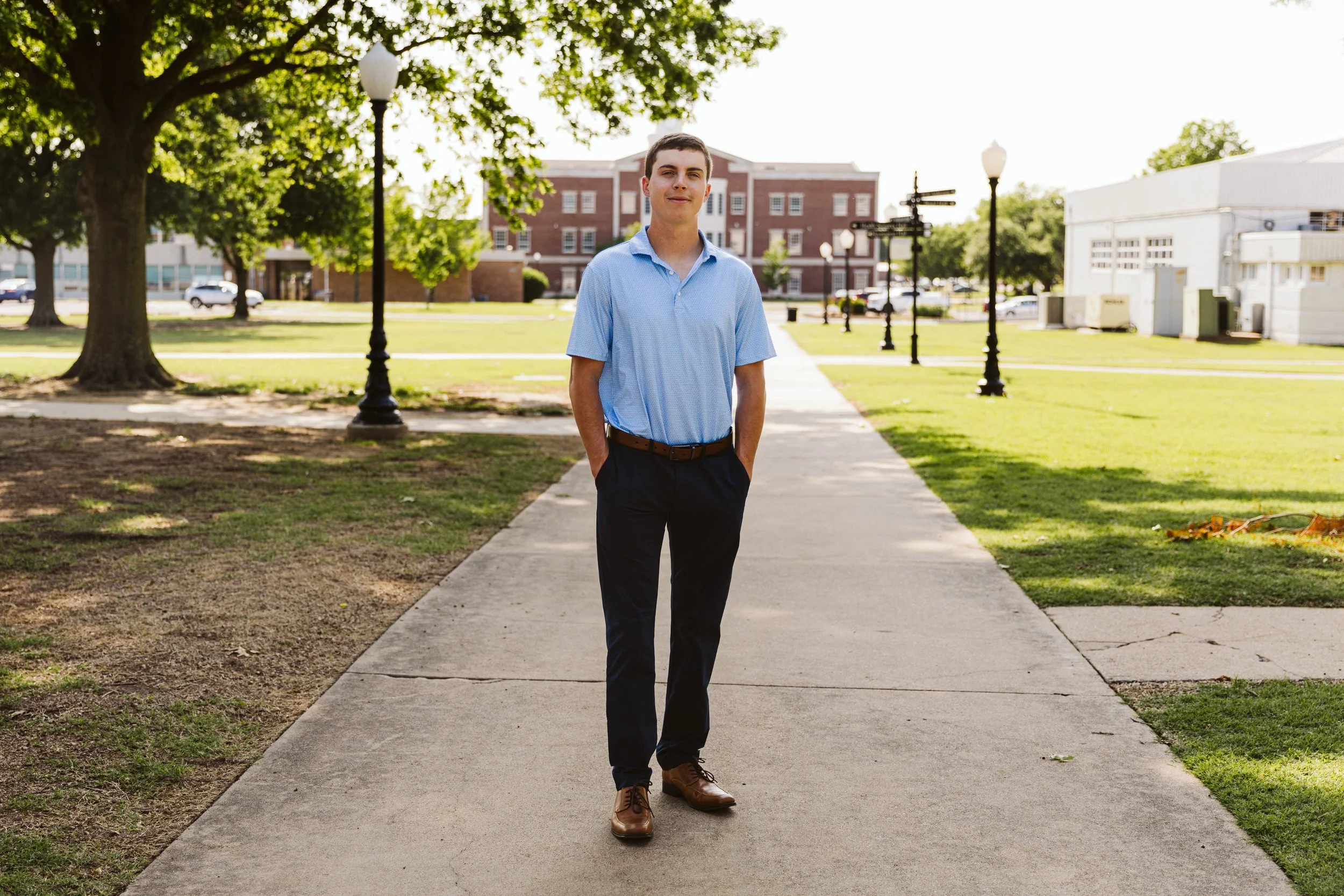 A person in a blue shirt and dark pants stands on a pathway in a park setting with trees and lampposts, a building in the background.