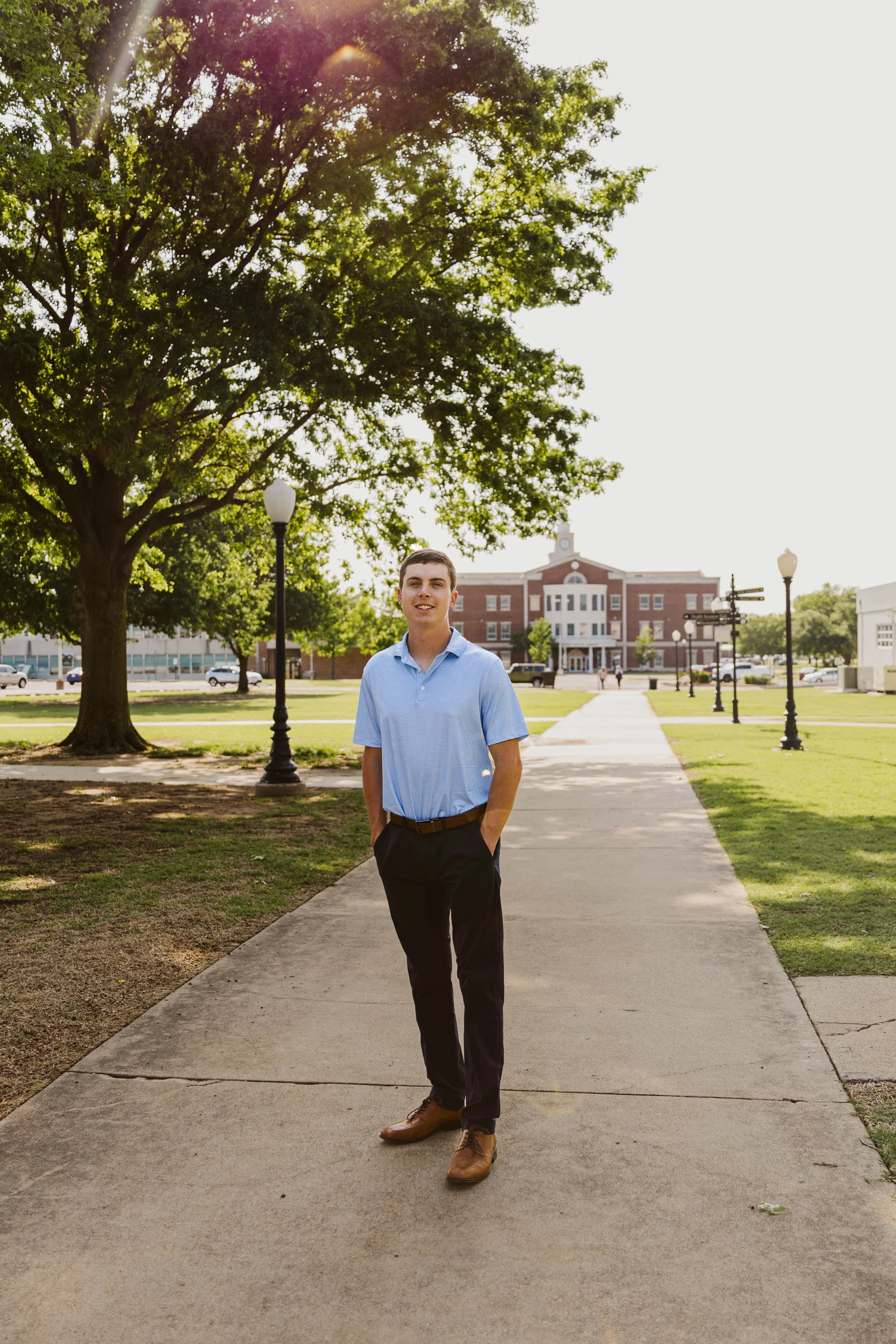 A person standing on a pathway in a park-like setting with trees, lampposts, and a large building in the background. The person is wearing a light blue shirt, dark pants, and brown shoes.