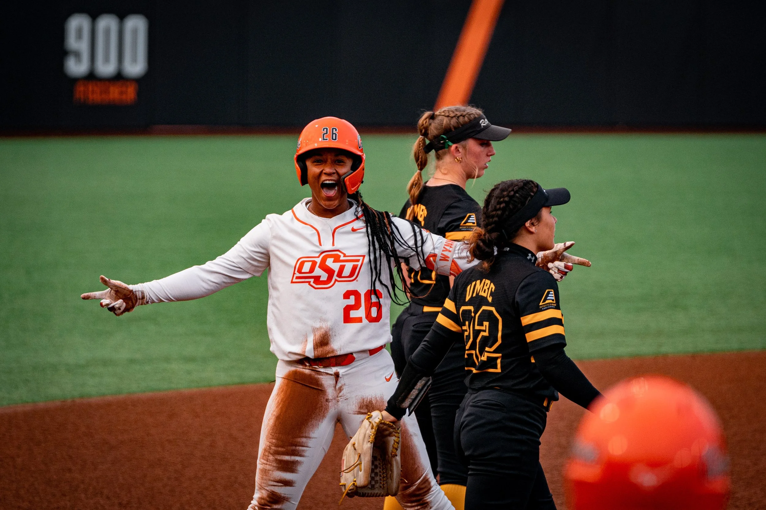 A softball player in a white and orange uniform celebrating on the field, surrounded by players in black and yellow uniforms.