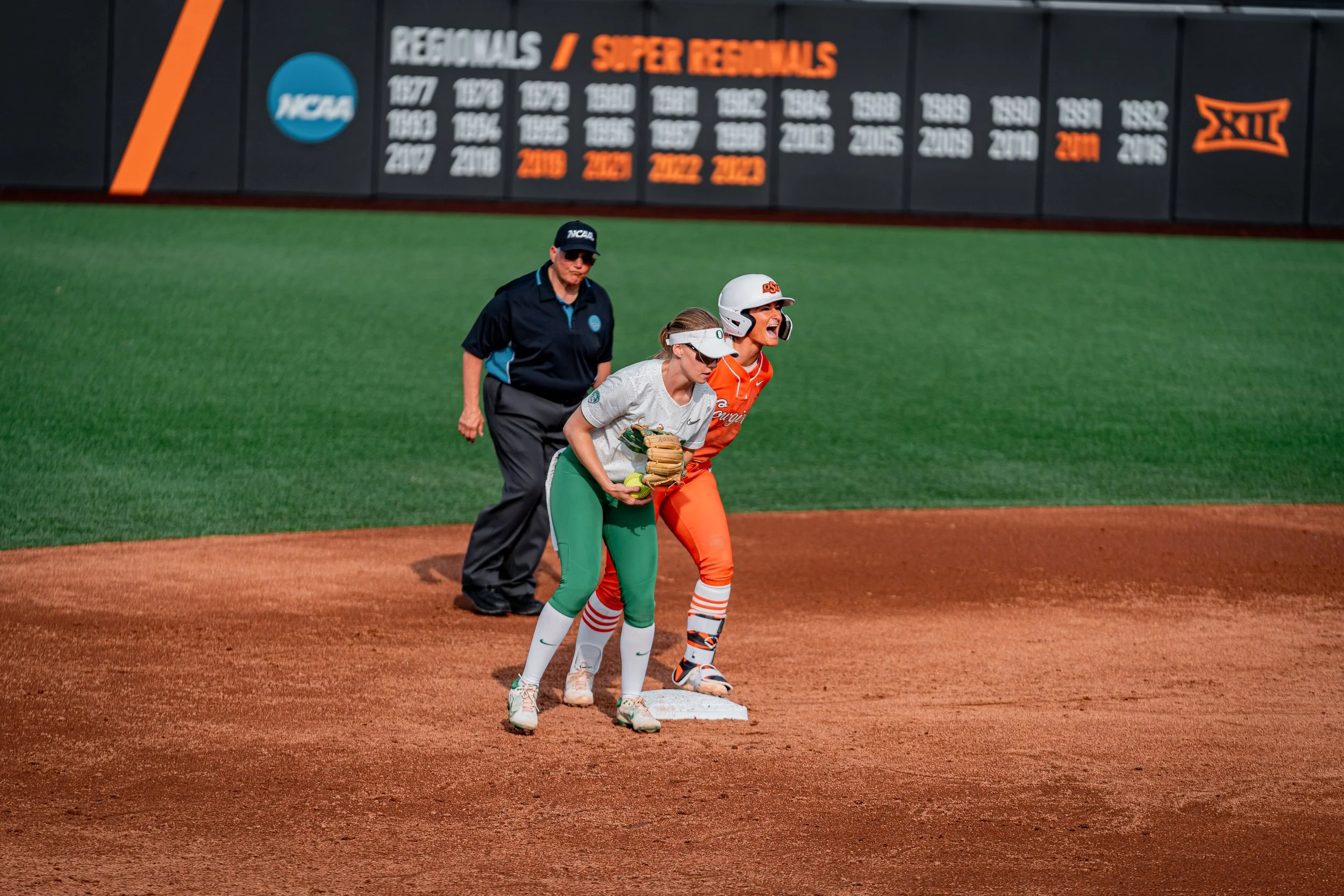 Softball players and umpire on the field during game with NCAA banner in background.