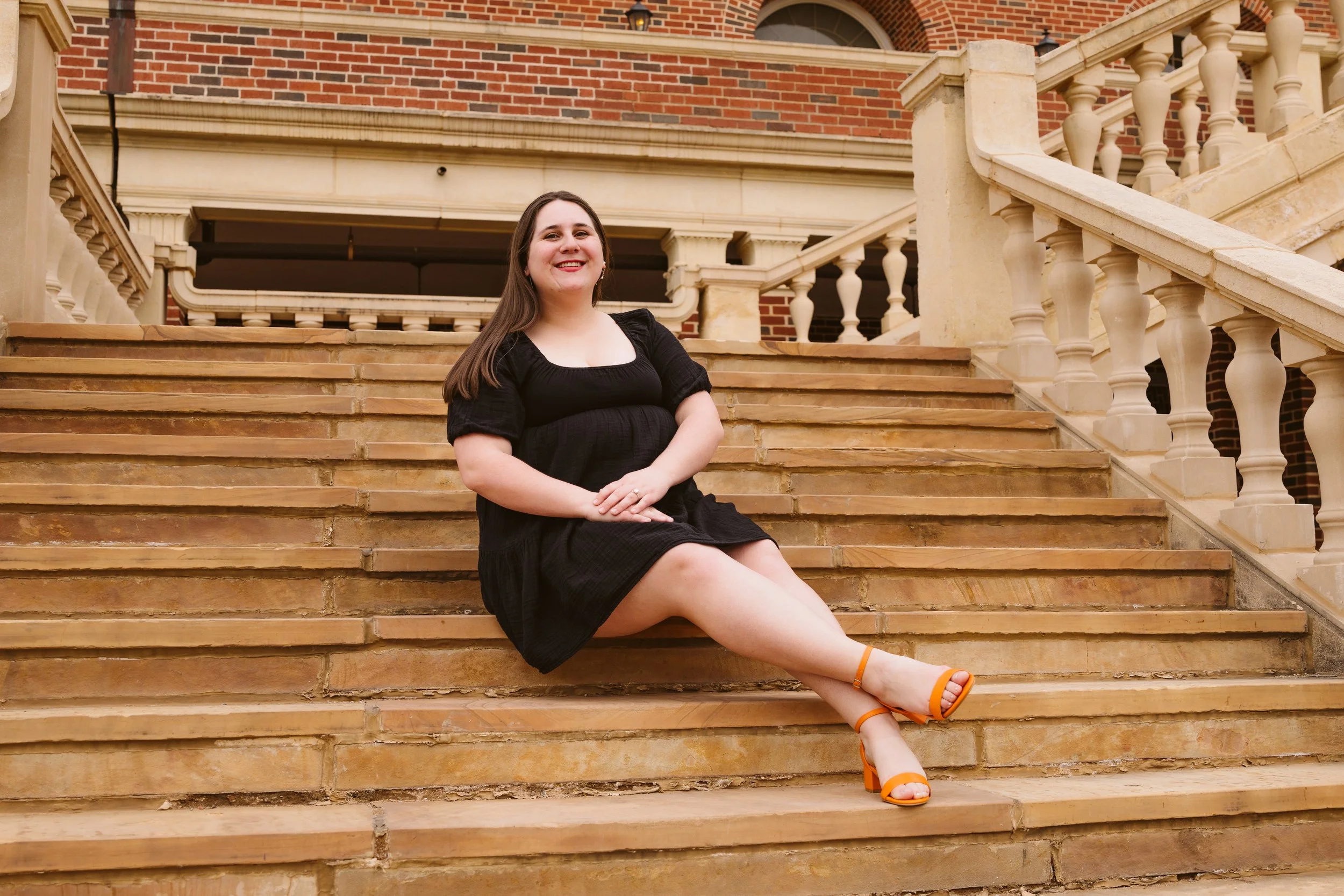 Woman seated on outdoor stone steps, wearing a black dress and orange shoes, with a brick building in the background.