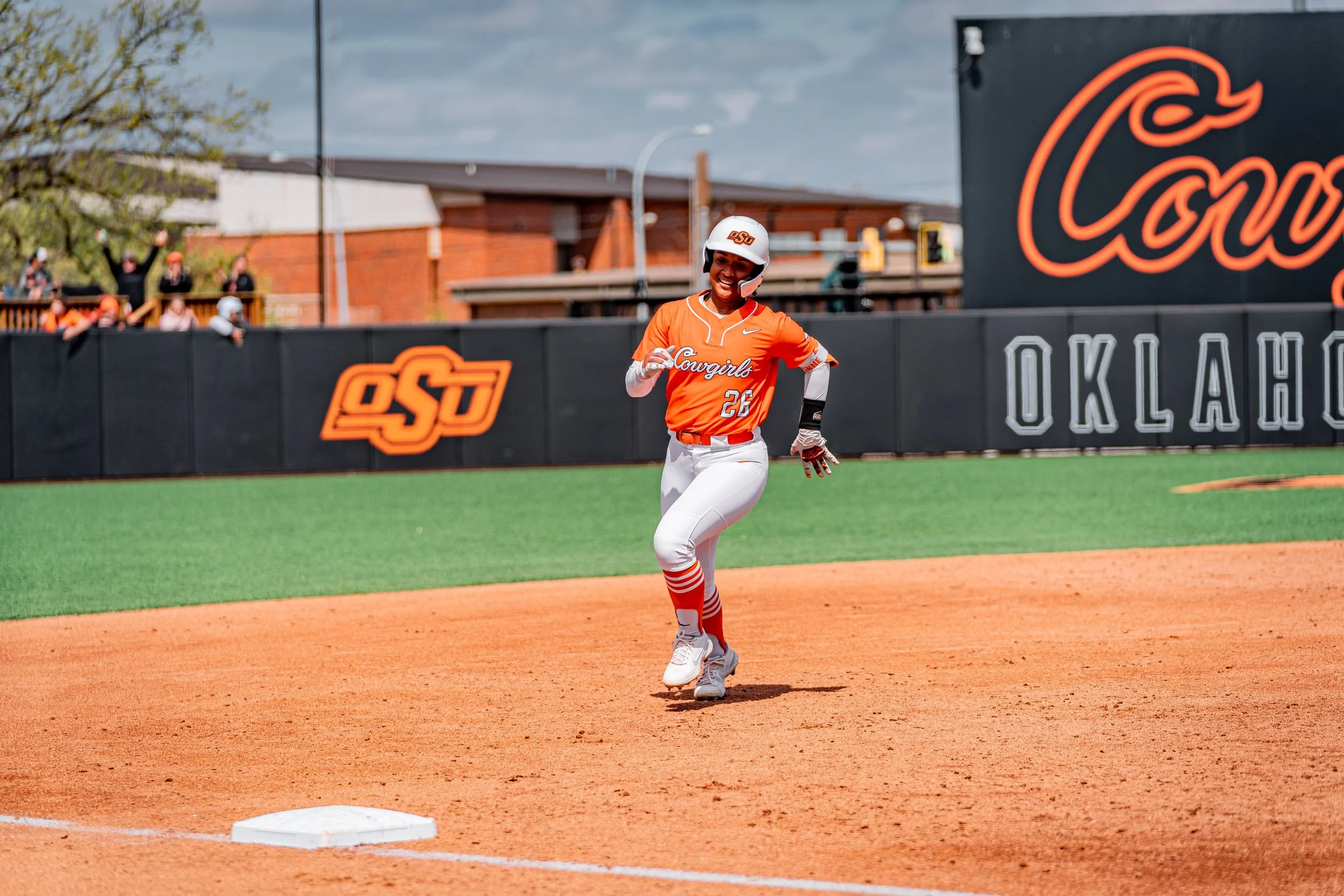 Softball player in orange uniform rounding a base on a field, with "OSU" and "Oklahoma" signage in the background.