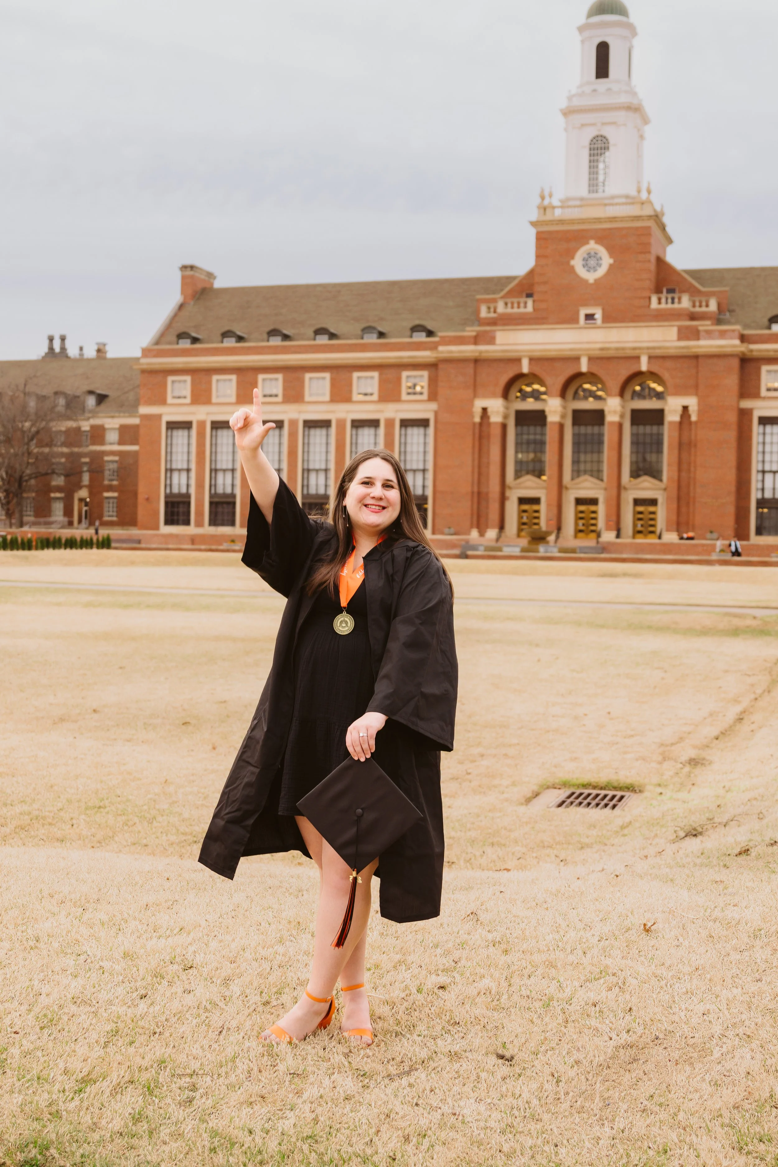 Graduate raising fist in celebration on campus with cap and gown, standing in front of a large brick building.