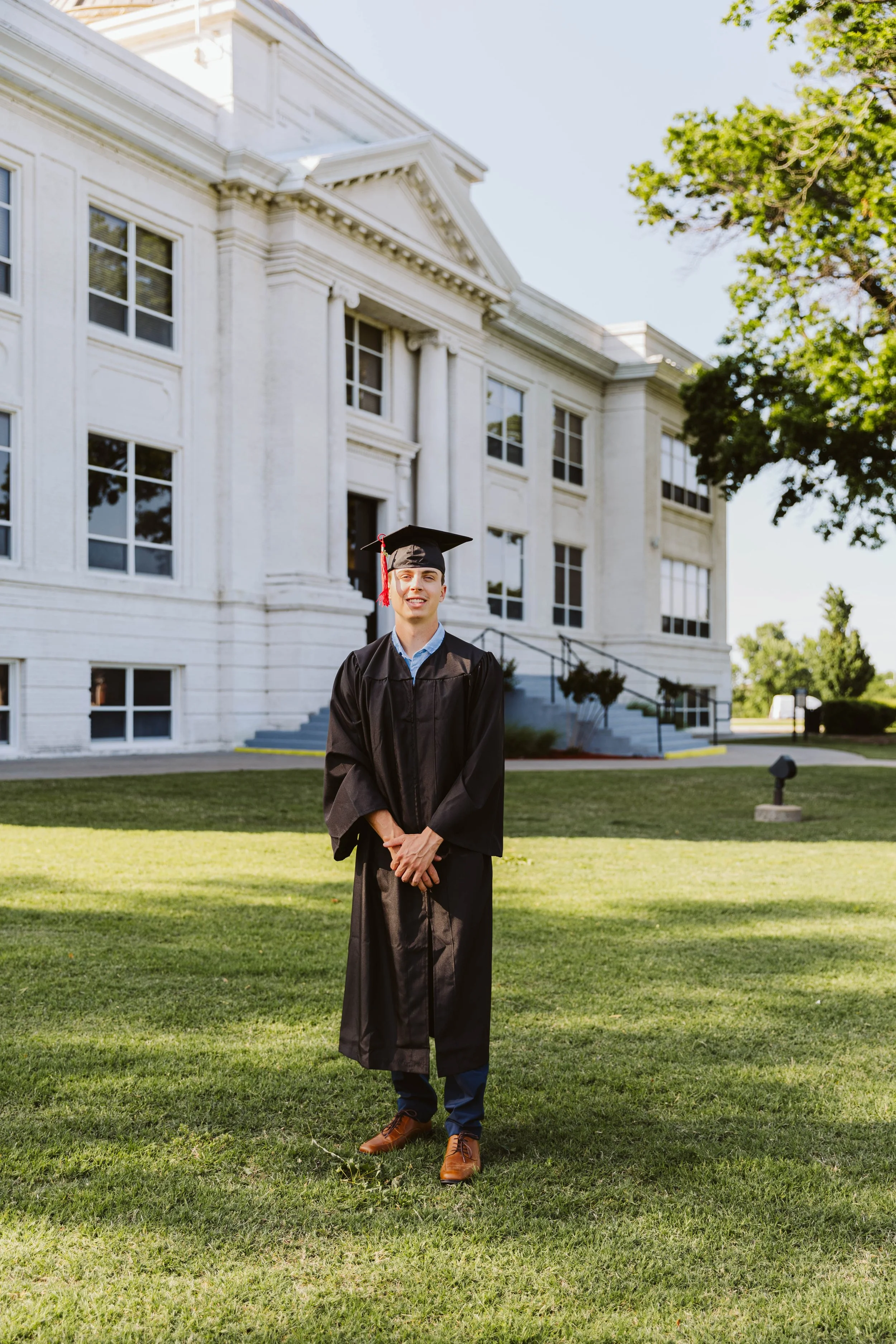 A person wearing a graduation cap and gown standing on grass in front of a white building.