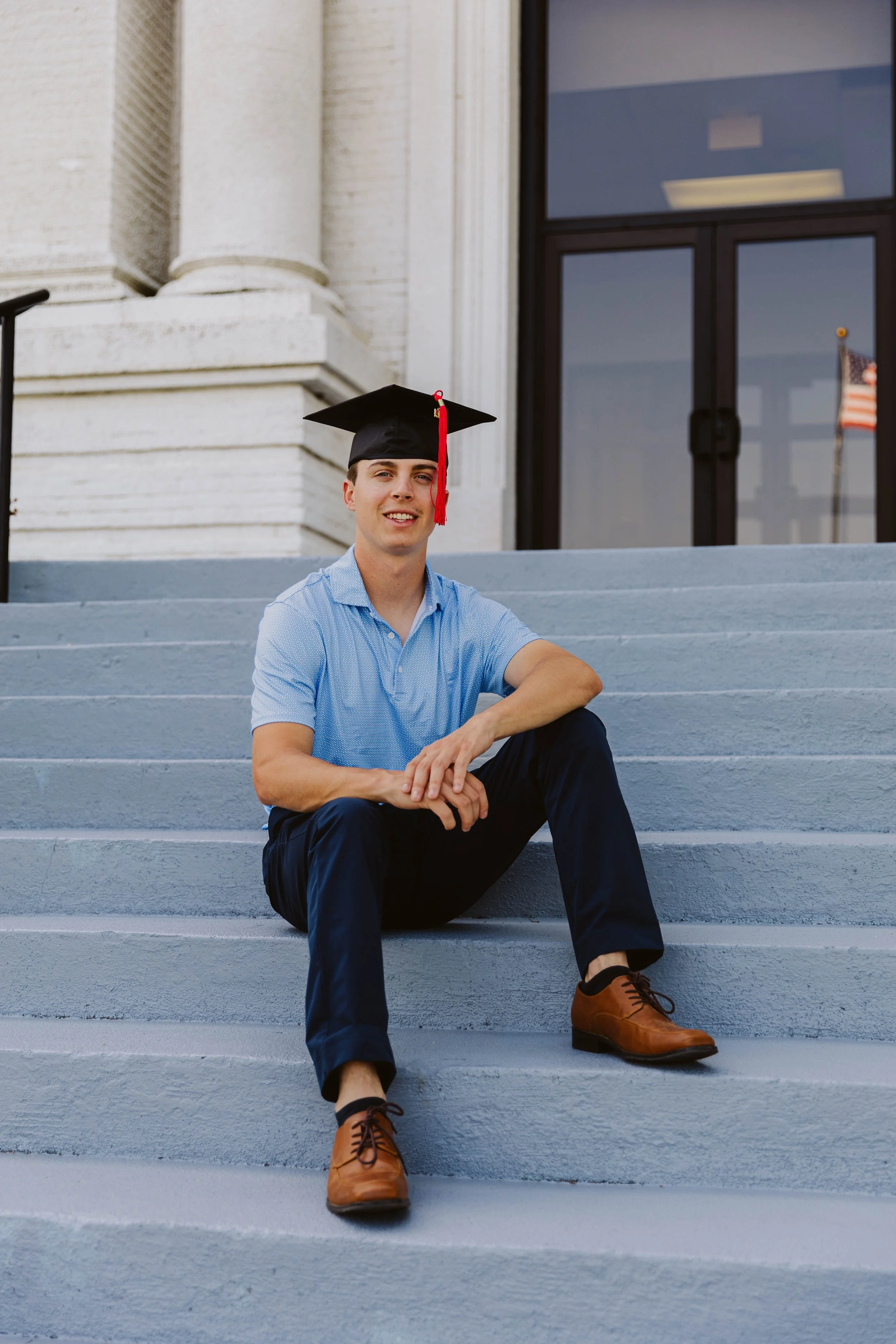 A young man wearing a graduation cap sits on the steps of a building with an American flag in the background.