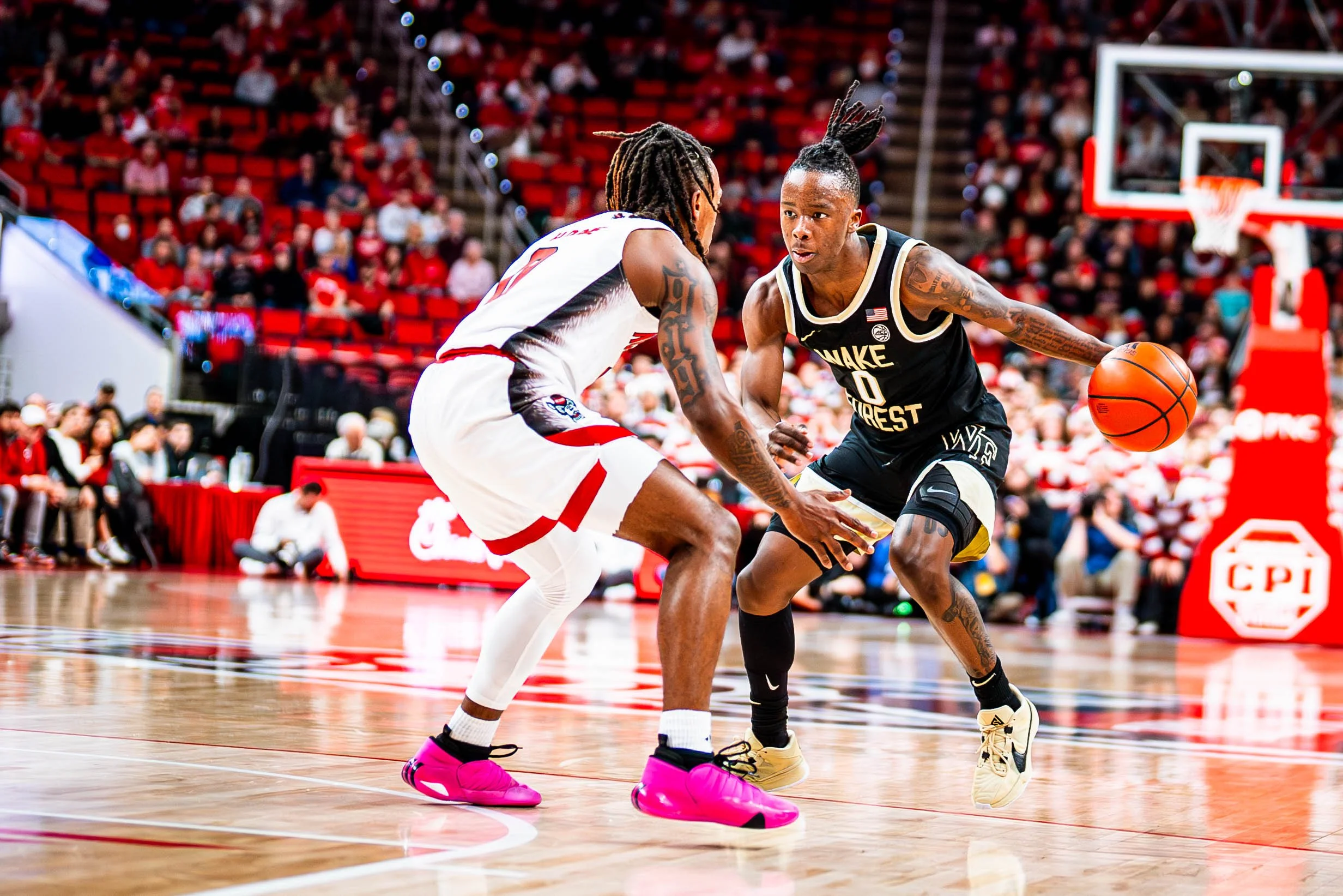 Two basketball players in action during a game, one wearing a black Wake Forest uniform and the other in a white jersey with red accents. The player in black is dribbling the ball while being closely defended.