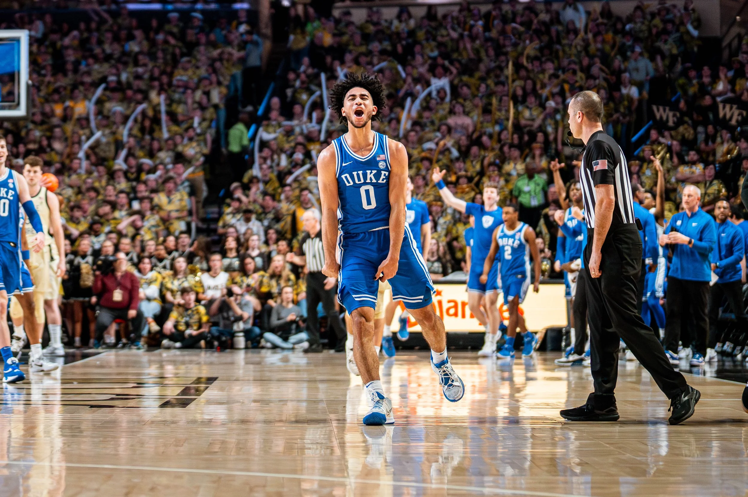 Basketball player in blue Duke uniform celebrating on court with cheering crowd and referee nearby.
