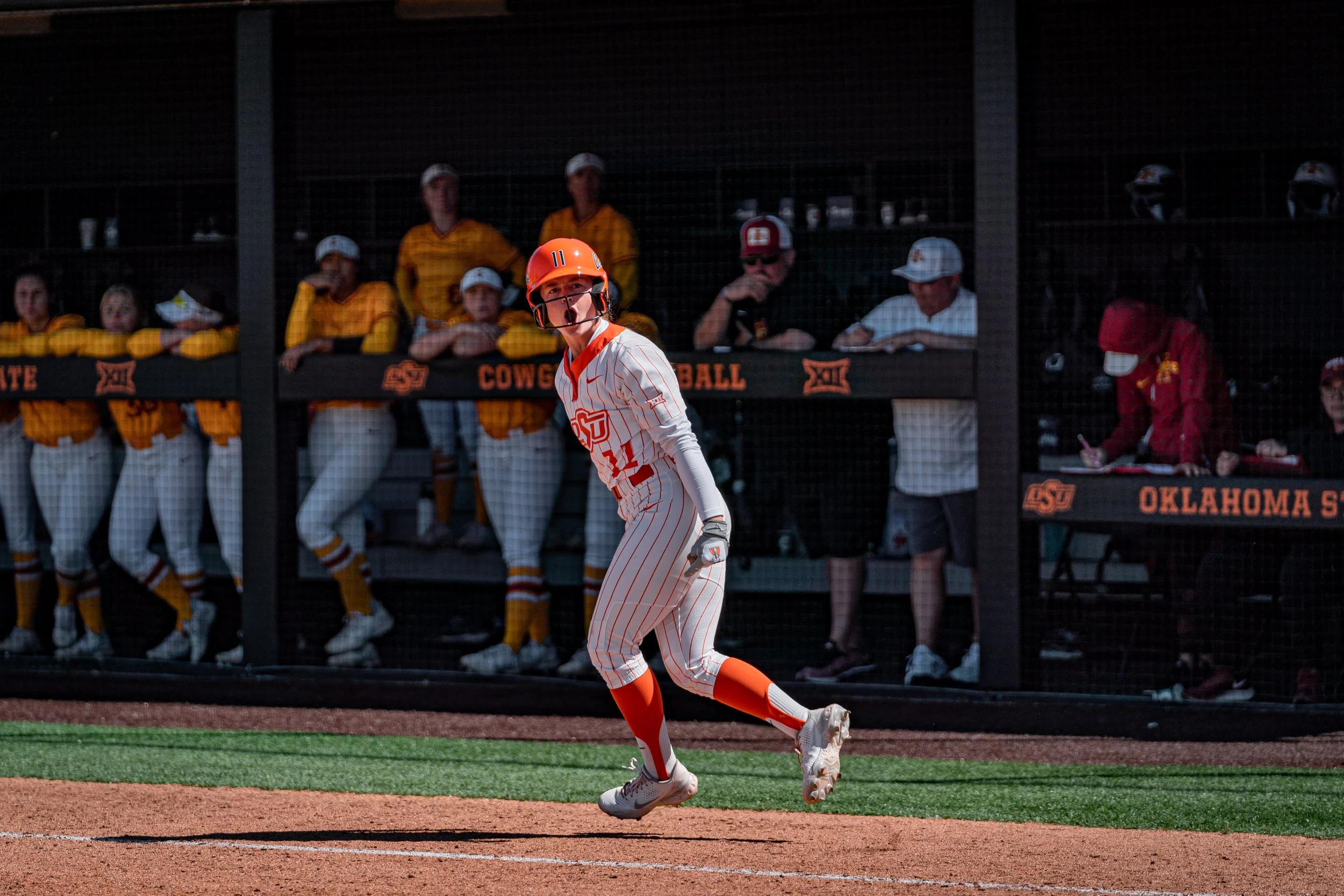 Softball player in orange and white uniform running near dugout during a game, with teammates and coaches watching from the bench.