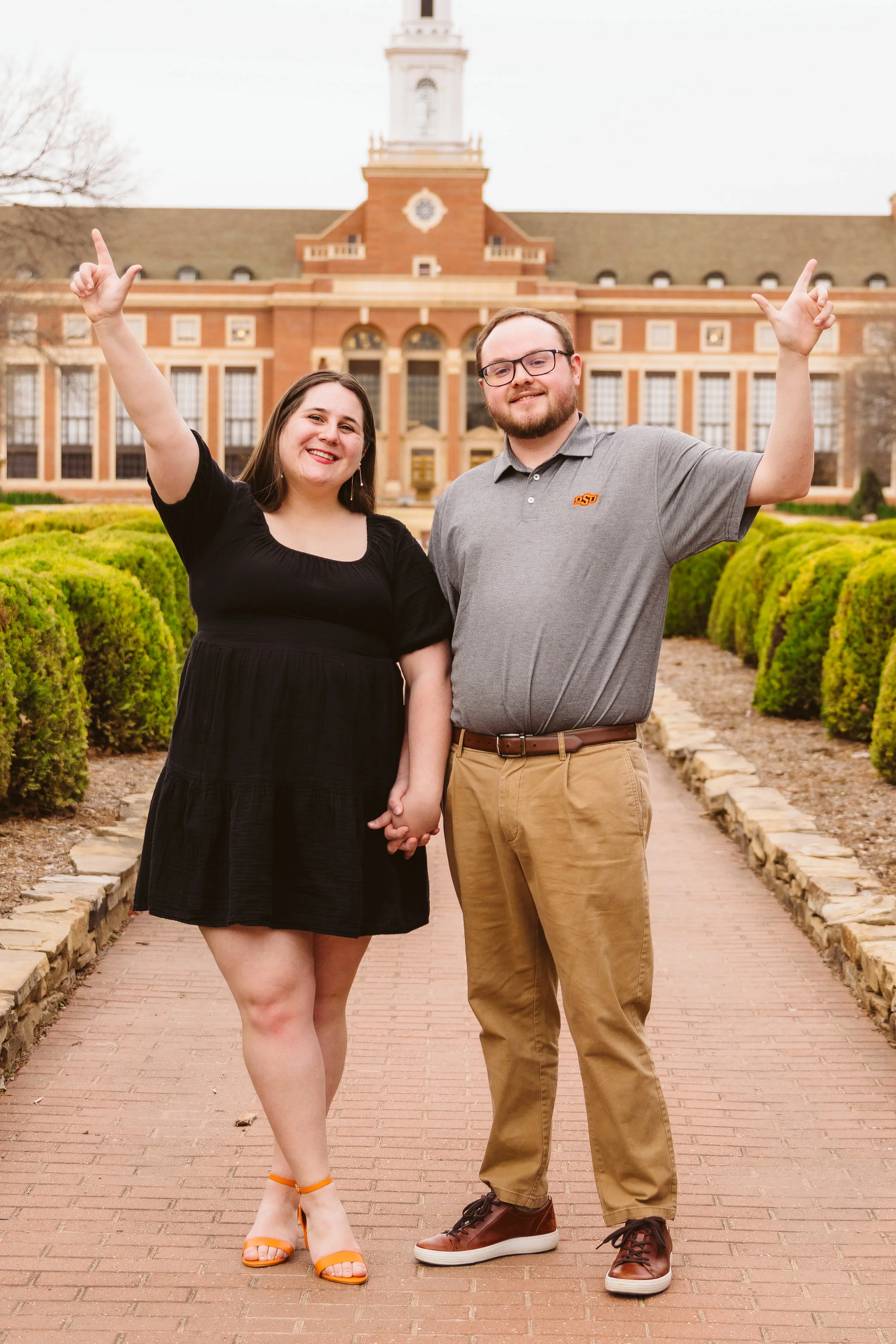 Two people standing outdoors on a brick pathway, holding hands, smiling, and raising one arm each. A historic brick building with a clock tower is in the background, surrounded by greenery.