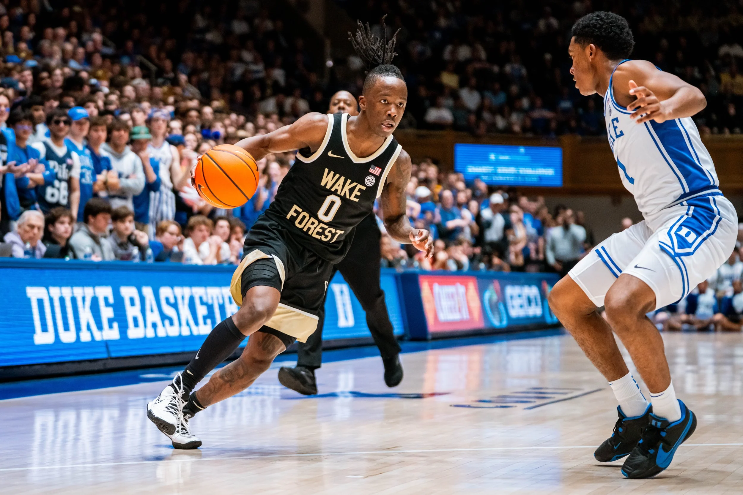 Basketball game between Wake Forest and Duke, with a Wake Forest player dribbling the ball past a Duke defender on the court. The audience is watching the action intensely.