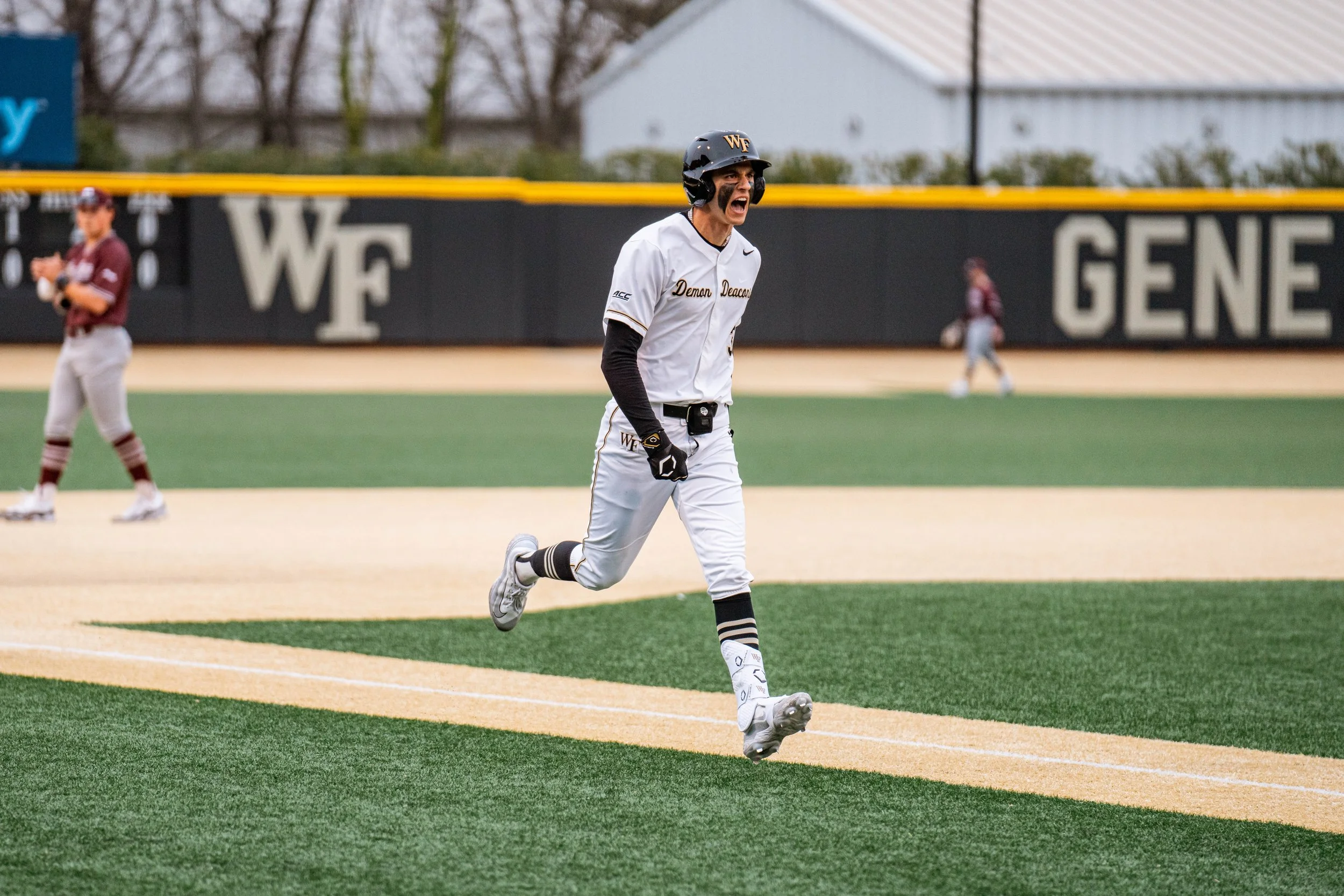 A baseball player in a white uniform and black helmet running on a field, showing excitement. The field has 'WF' and 'GENE' written in the background, with trees and a building visible.