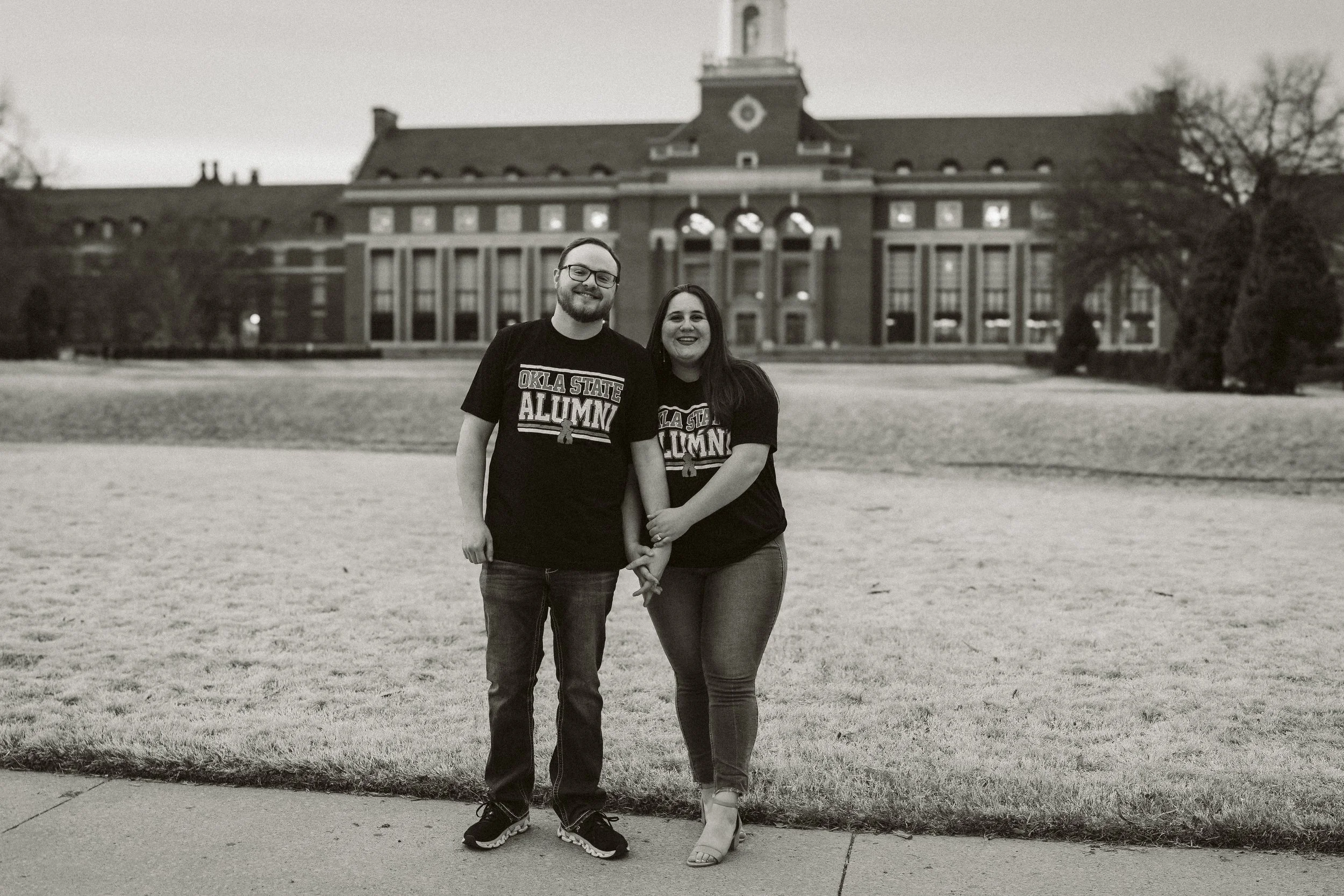 Two individuals wearing matching "Okla State Alumni" T-shirts stand in front of a large building on a grassy lawn, posing for the camera with linked hands.