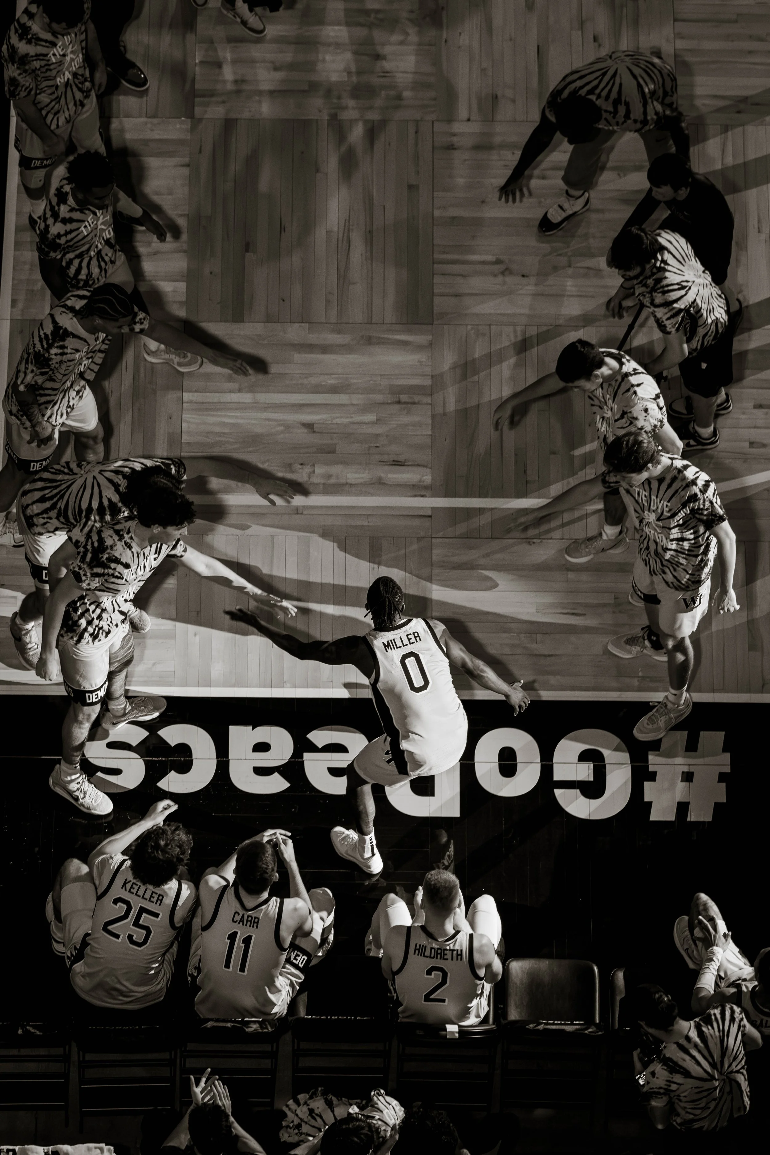 Basketball player high-fiving teammates on the court, with players seated on the bench. Hashtag and court markings visible.