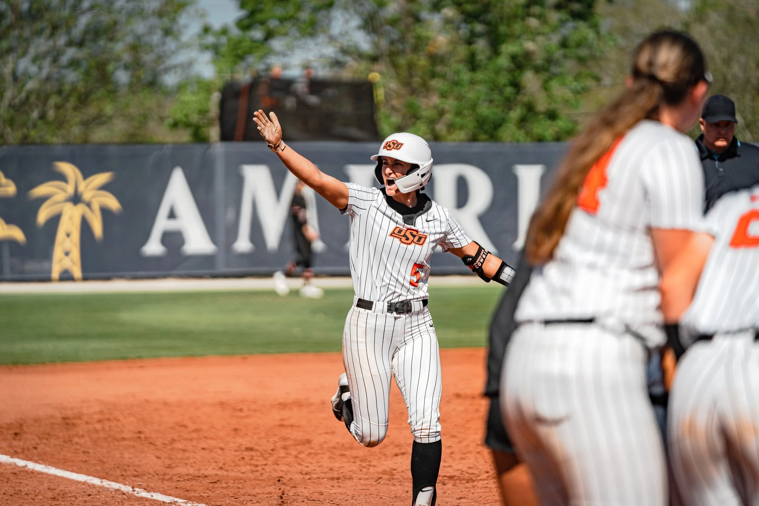 Softball player celebrating after scoring, wearing white pinstriped uniform with "OSU" emblem, running on a field with a palm tree backdrop.