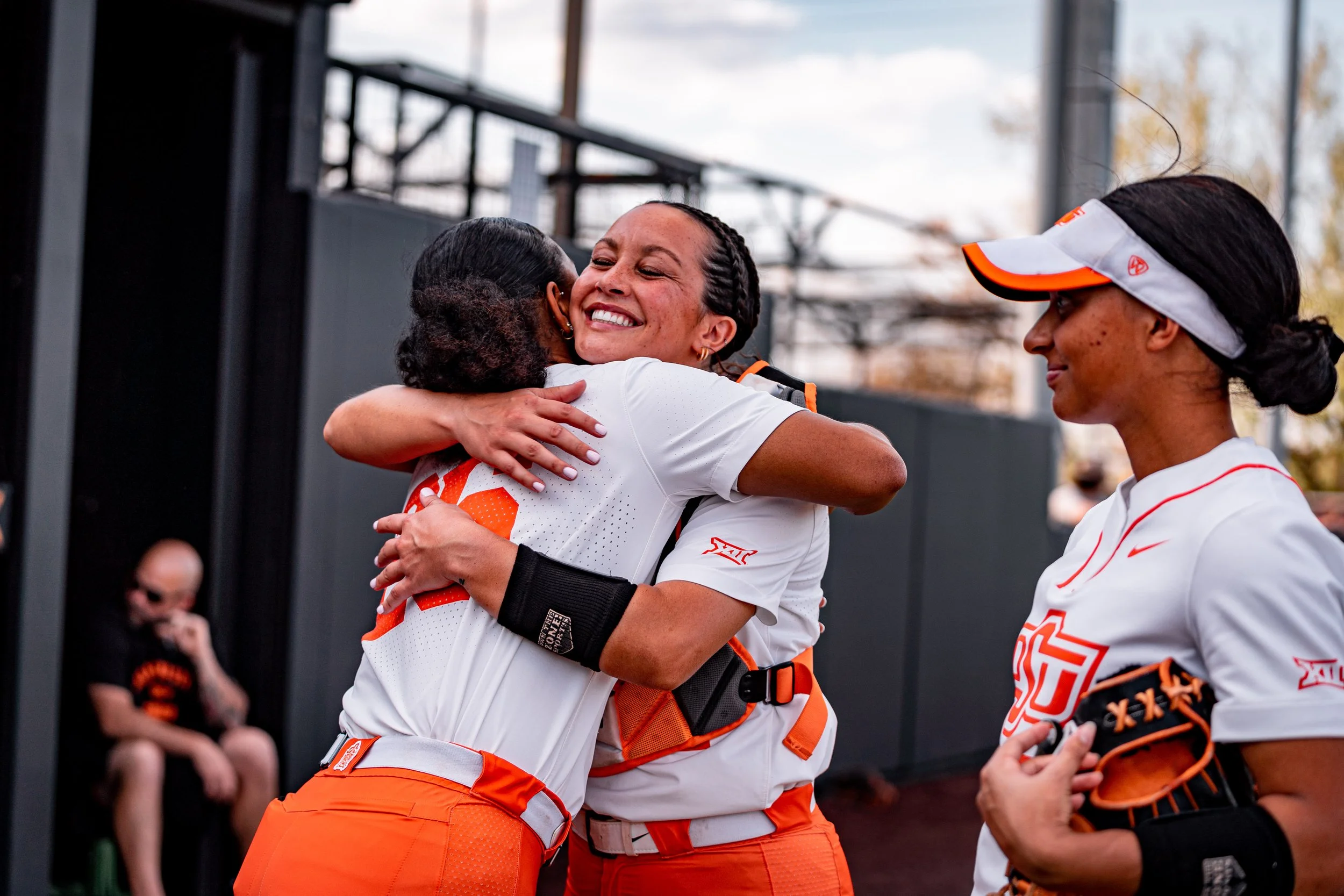 Two female softball players in orange and white uniforms hugging and smiling, with a third player standing nearby wearing a white visor. They are outdoors near a dugout.