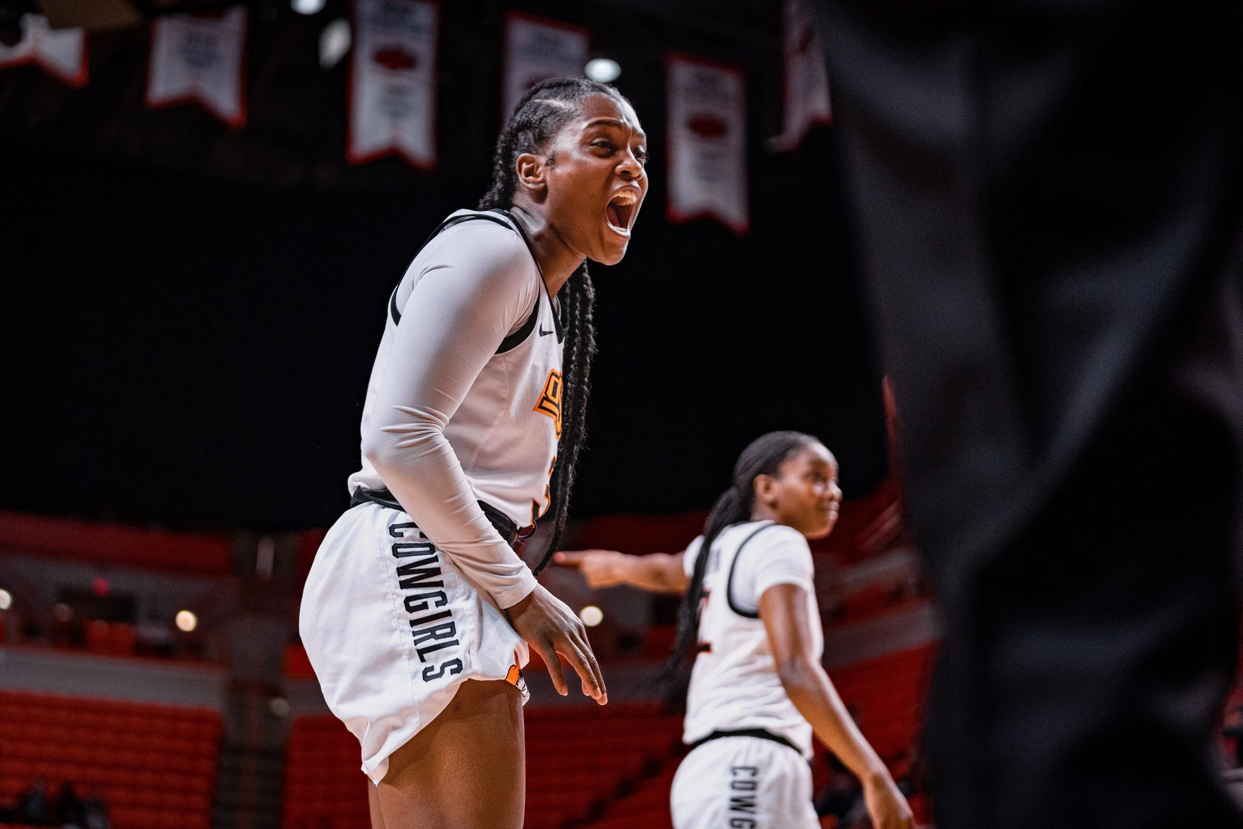 Female basketball player in white uniform with 'Cowgirls' printed on shorts, shouting during a game, basketball court setting.